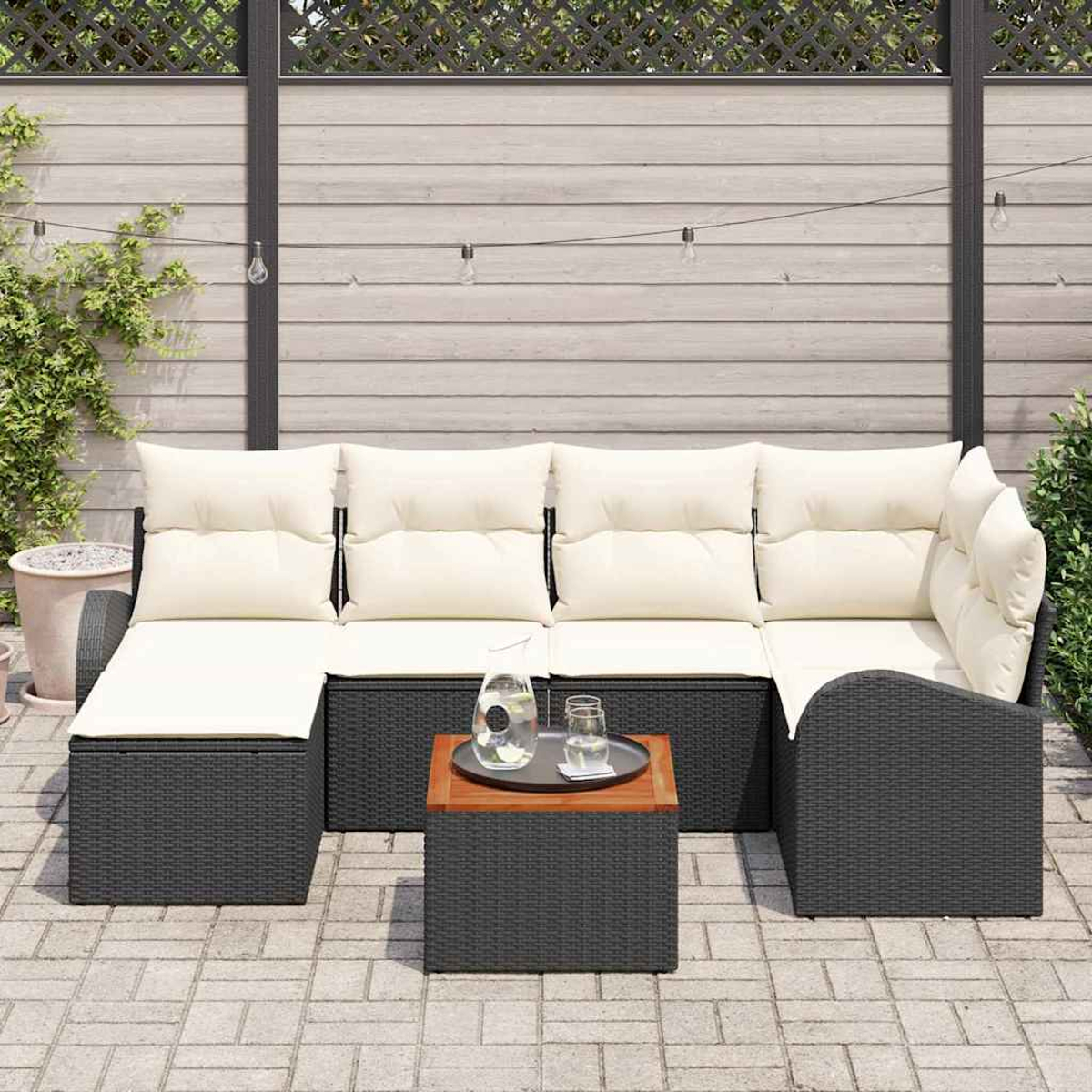 Sofá seccional exterior con cojines crema, mesa de madera con jarra y vasos de vidrio, contra una valla de madera.