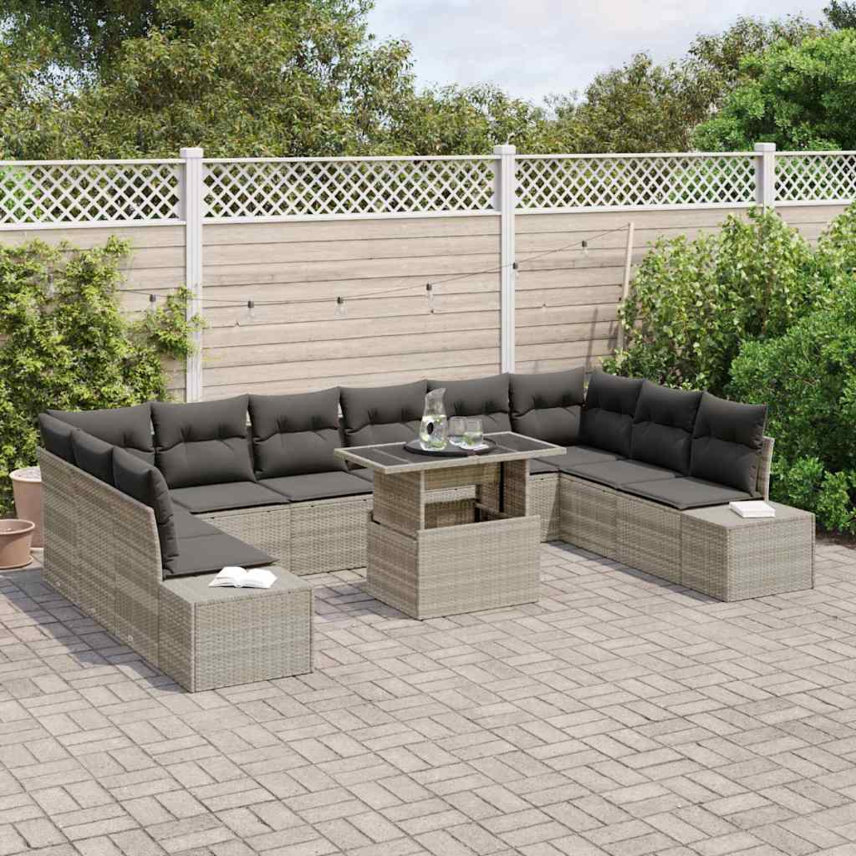 Eine Terrassengarnitur mit grauen Kissen, einem Tisch mit Getränken, einer grauen Ziegelterrasse und einem weißen Zaun mit Grün.