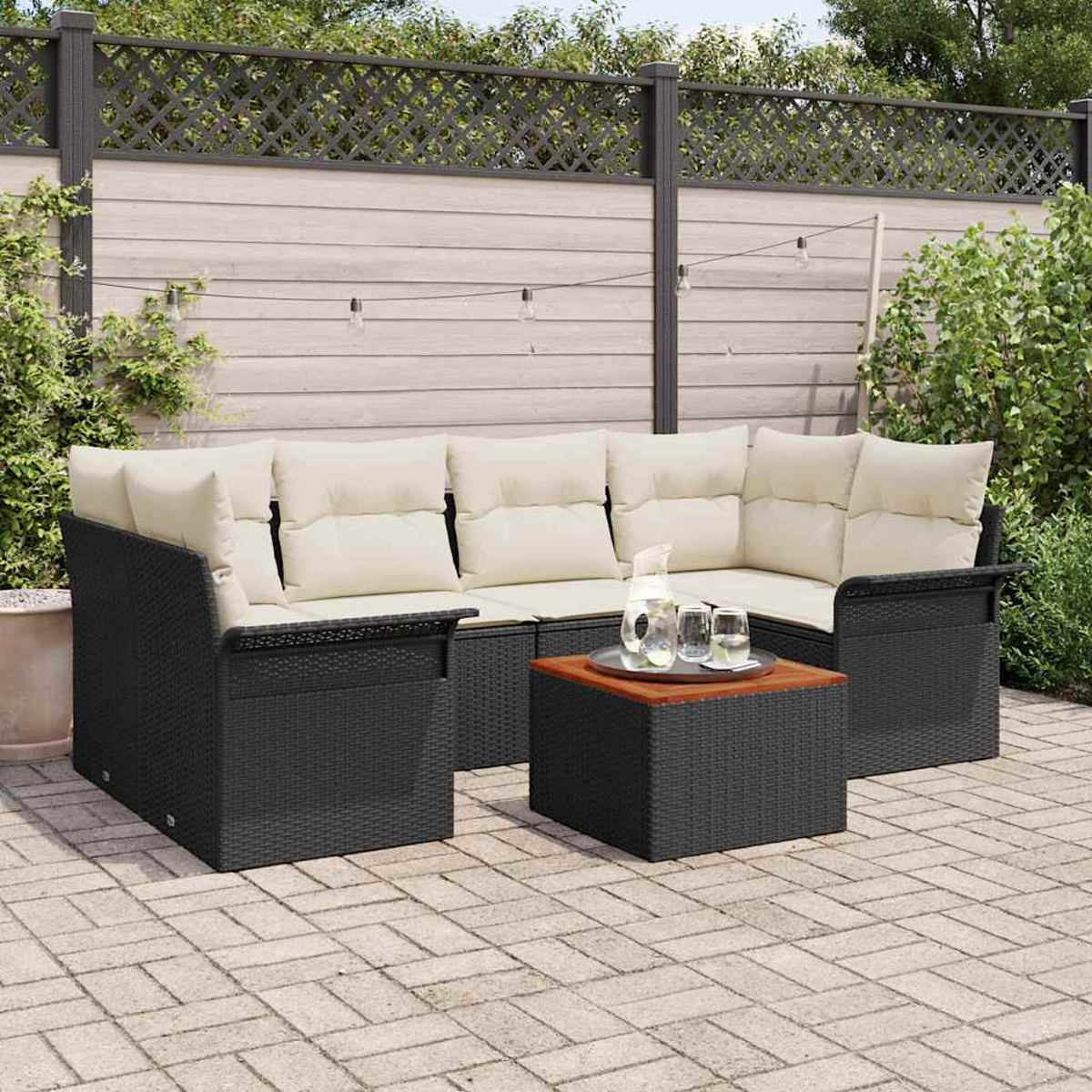 Garten-Sitzgruppe mit schwarzen Korbflechtrahmen und cremefarbenen Kissen, einem kleinen Tisch mit Holzplatte und einem Krug mit Gläsern auf einer Terrasse.