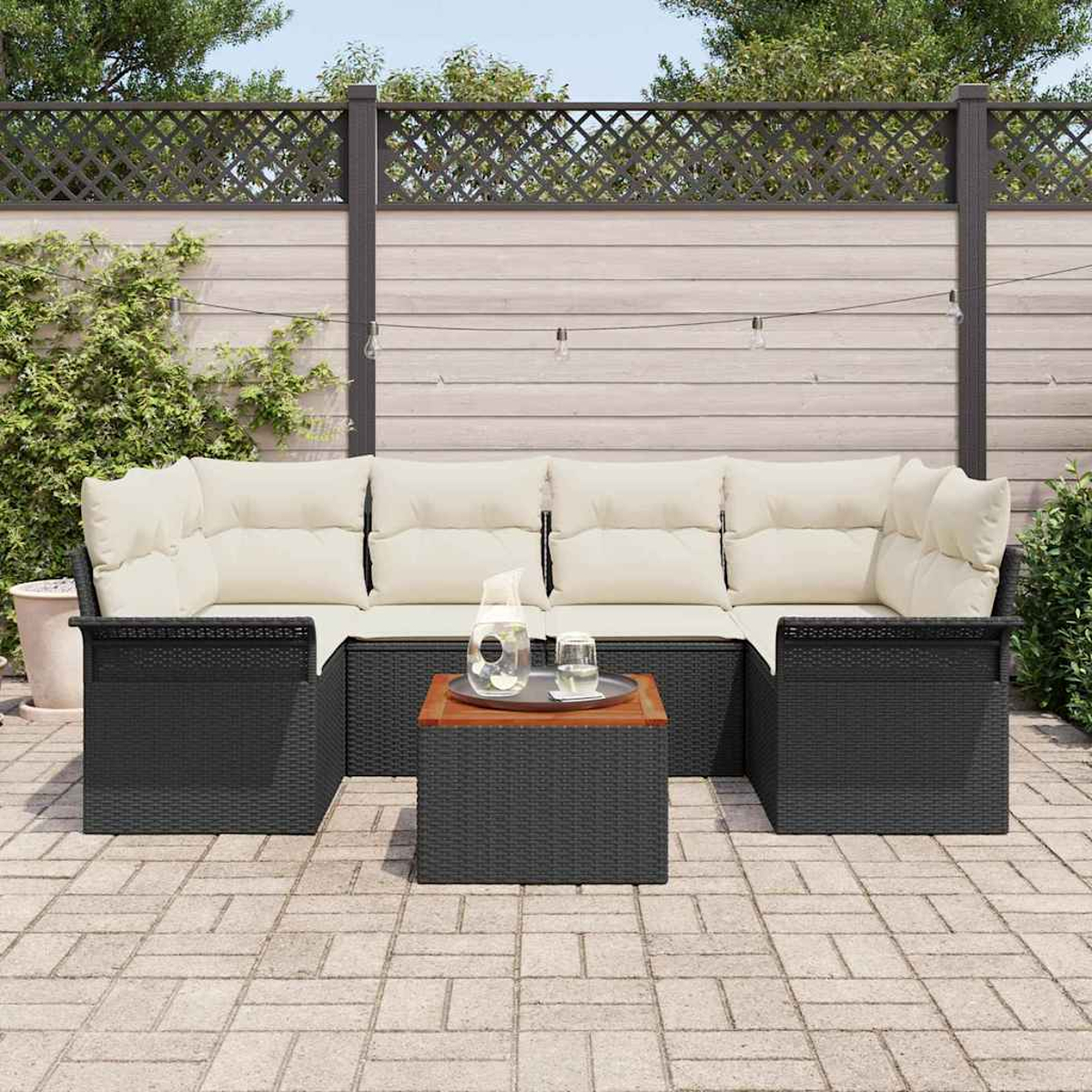 Terrassenset mit schwarzer Korbmöbel, cremefarbenen Kissen und einem Holztisch. Ein Zaun und Grün im Hintergrund.