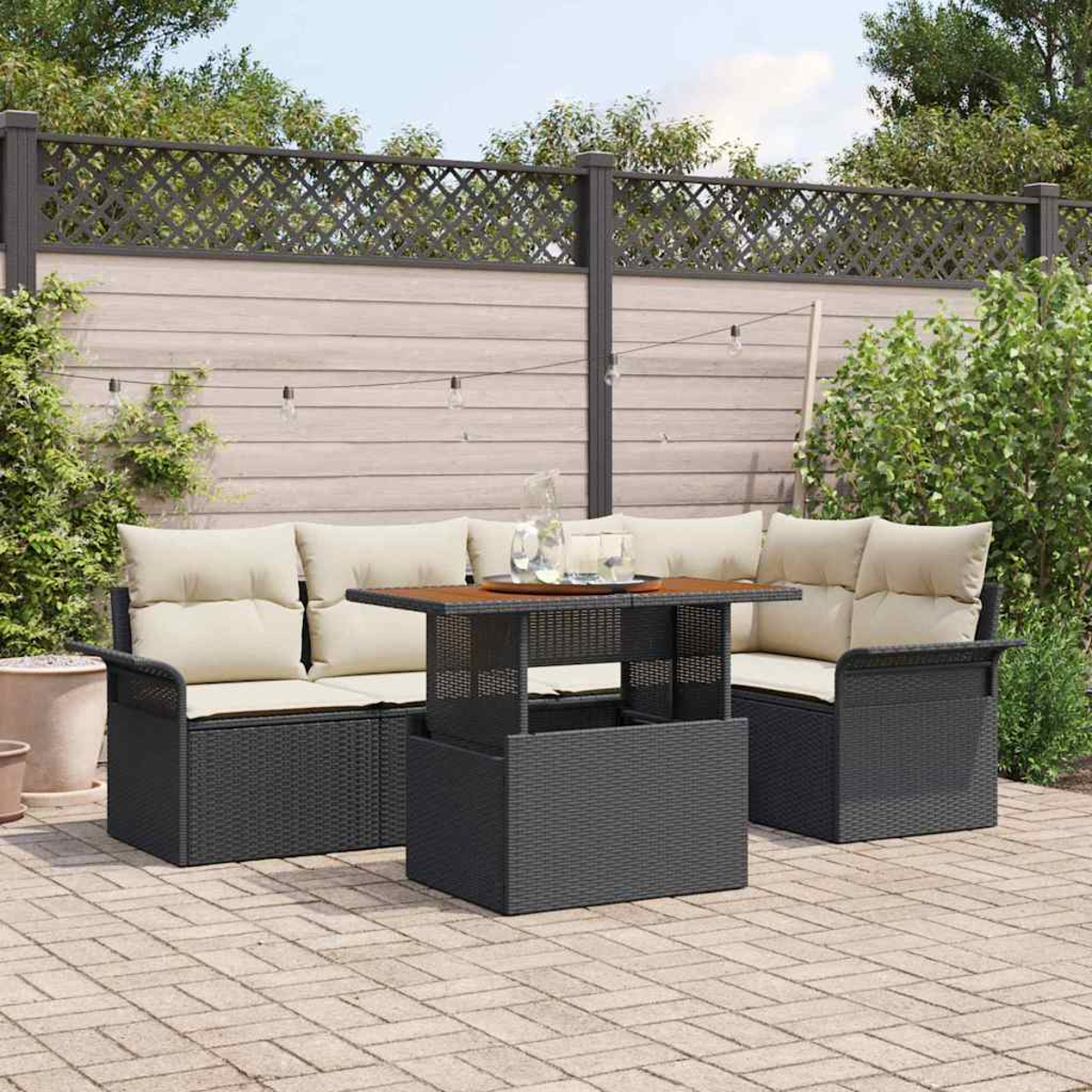 Garten-Terrassenset mit schwarzen Korbmöbeln und beigefarbenen Kissen. Ein Holztisch mit Getränken steht im Zentrum. Ein Zaun und Pflanzen sind im Hintergrund.