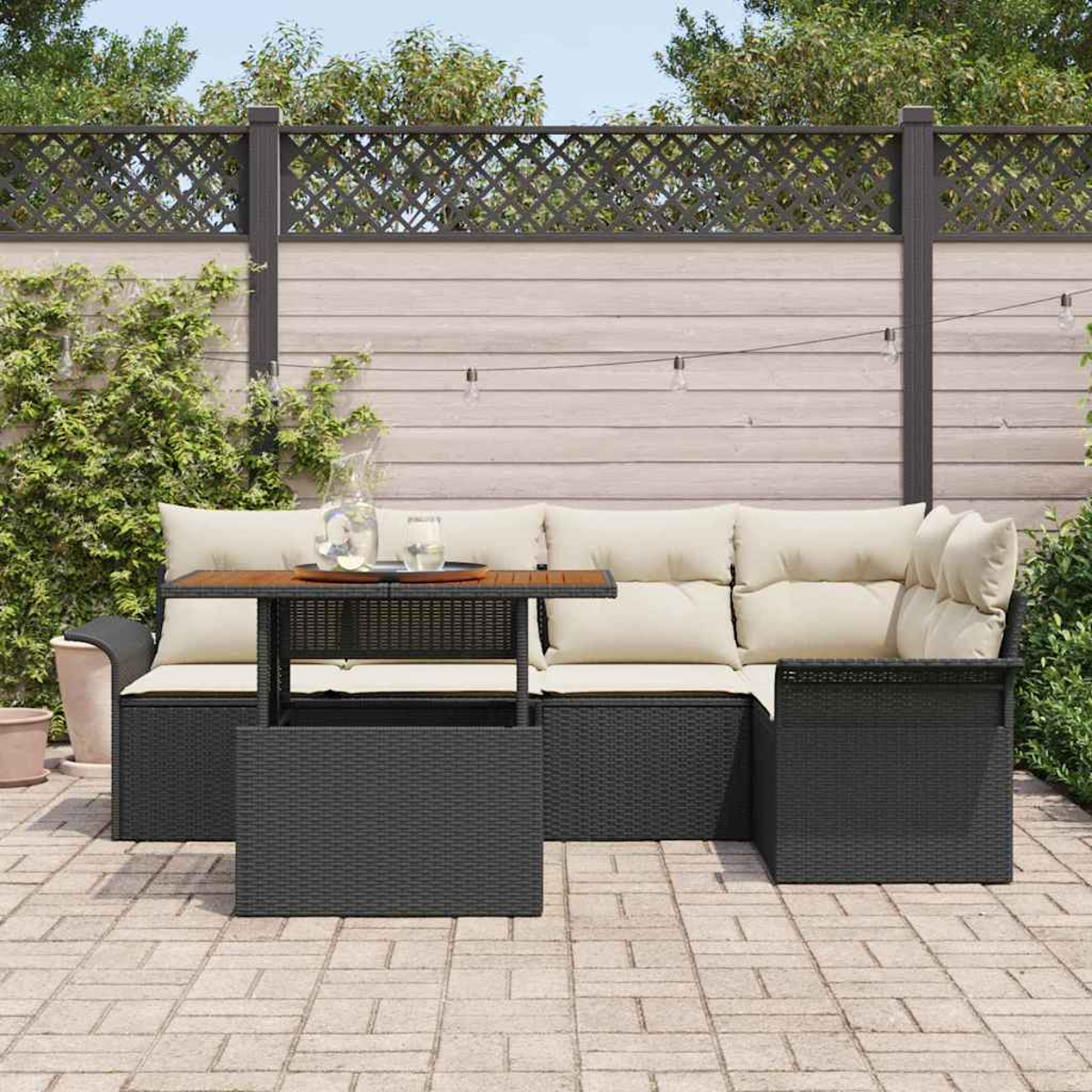 Garten-Terrassenset mit schwarzer Korbmöbel, beigen Kissen und einem Holztisch. Hintergrund ist ein Zaun und Grün.