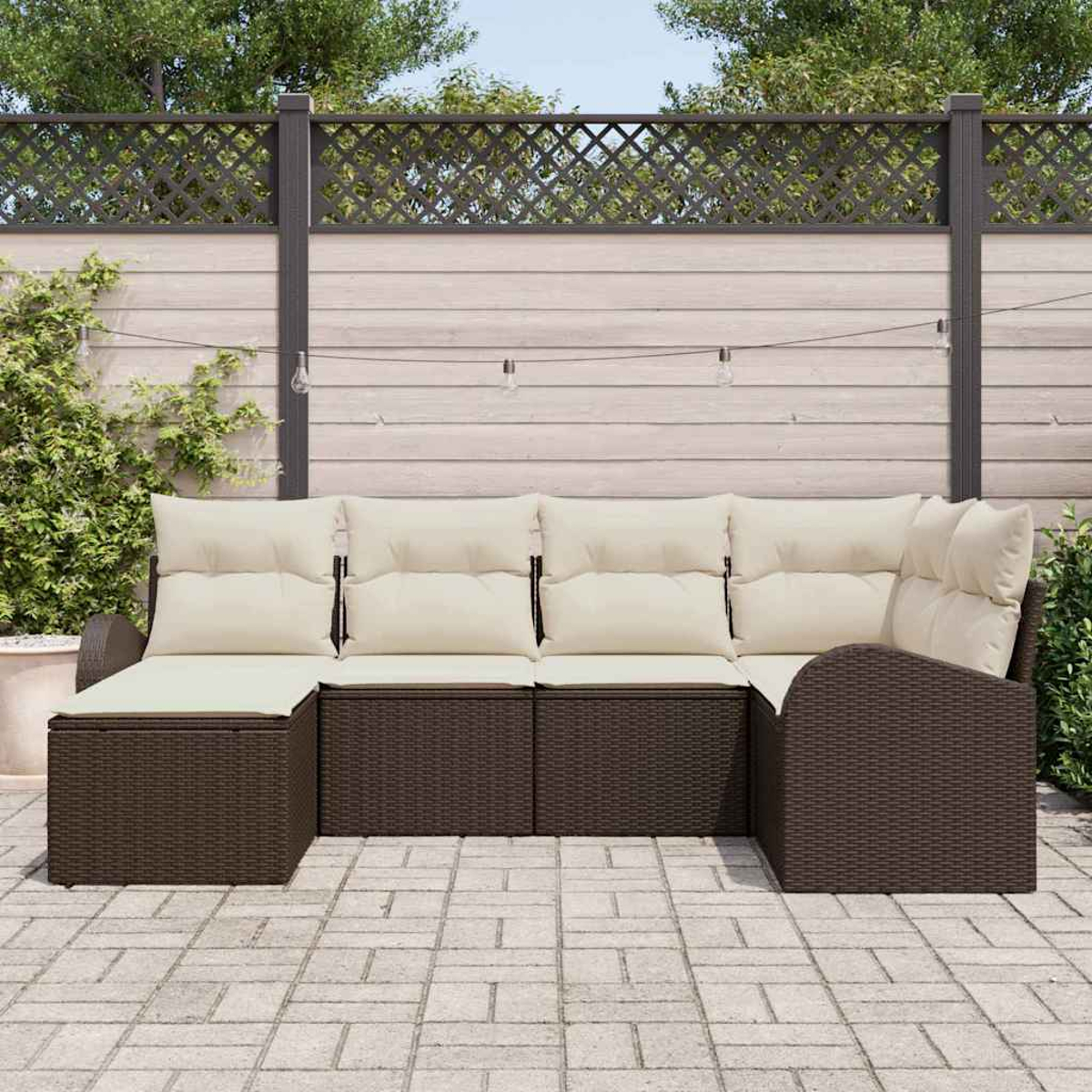 Braunes Outdoor-Wicker-Ecksofa mit cremefarbenen Kissen auf einer Stein-Terrasse. Ein Holzzaun und Grünzeug sind im Hintergrund.