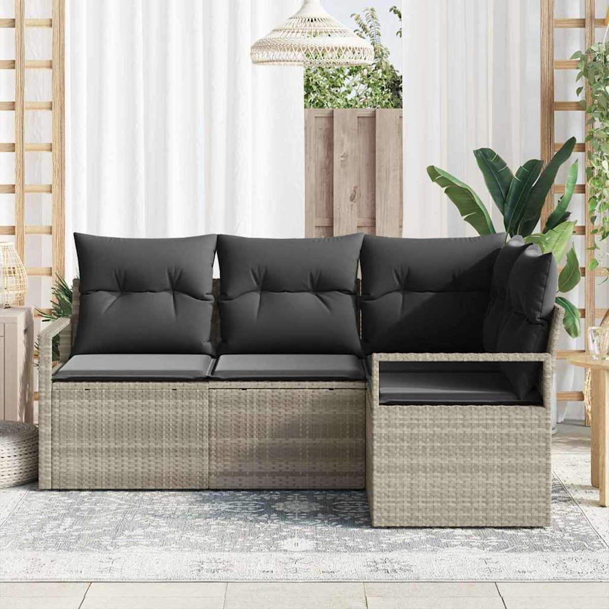 Ein graues Outdoor-Ecksofa aus Korbgeflecht mit dunkelgrauen Kissen steht auf einem gemusterten Teppich. Weiße Vorhänge und Pflanzen sind im Hintergrund.