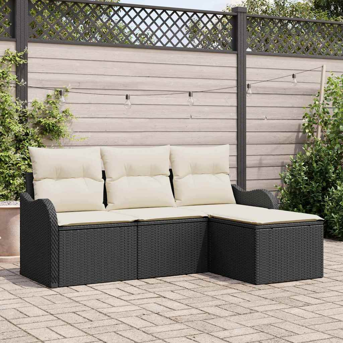 Ein schwarzes Korbsessel-Ecksofa mit cremefarbenen Kissen steht auf einer Backsteinterrasse. Ein Holzzaun und Pflanzen sind im Hintergrund.