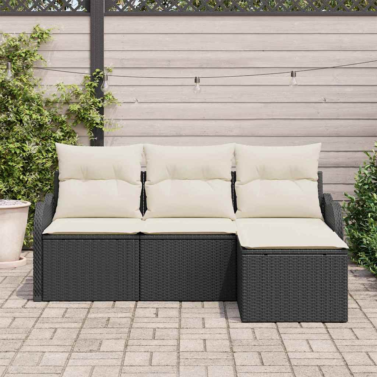 Outdoor-Ecksofa mit hellen Kissen und dunklem Korbgeweberahmen. Vor einem Holzzaun mit Lichterketten und Grün.