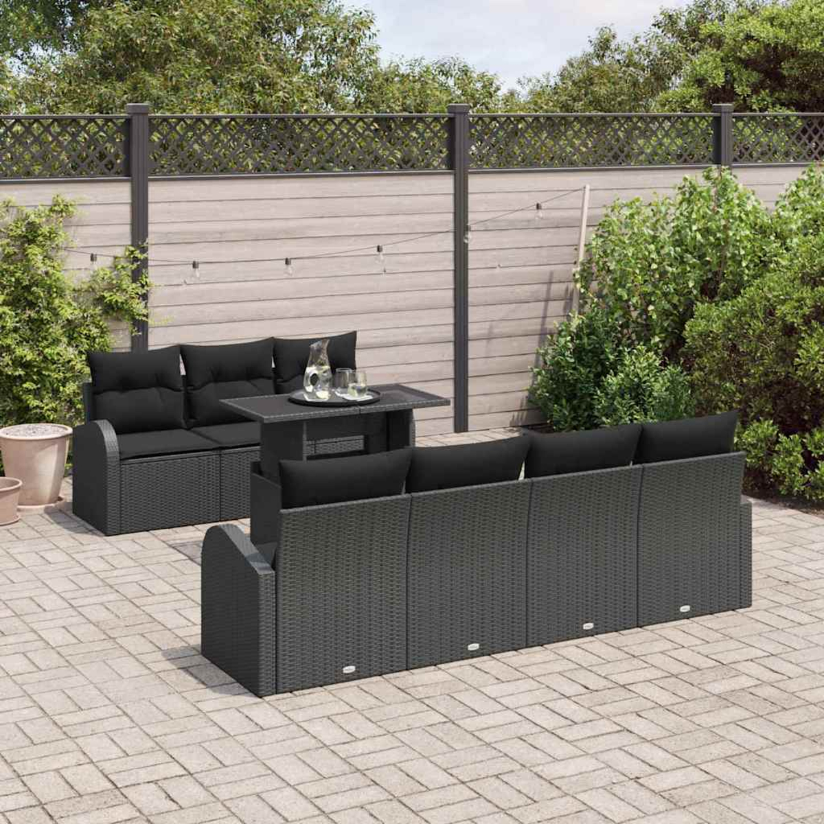 Garten-Set mit schwarzen Kissen, einem Tisch und grauem Korbmuster auf einer Steinterrasse, mit Zaun im Hintergrund.