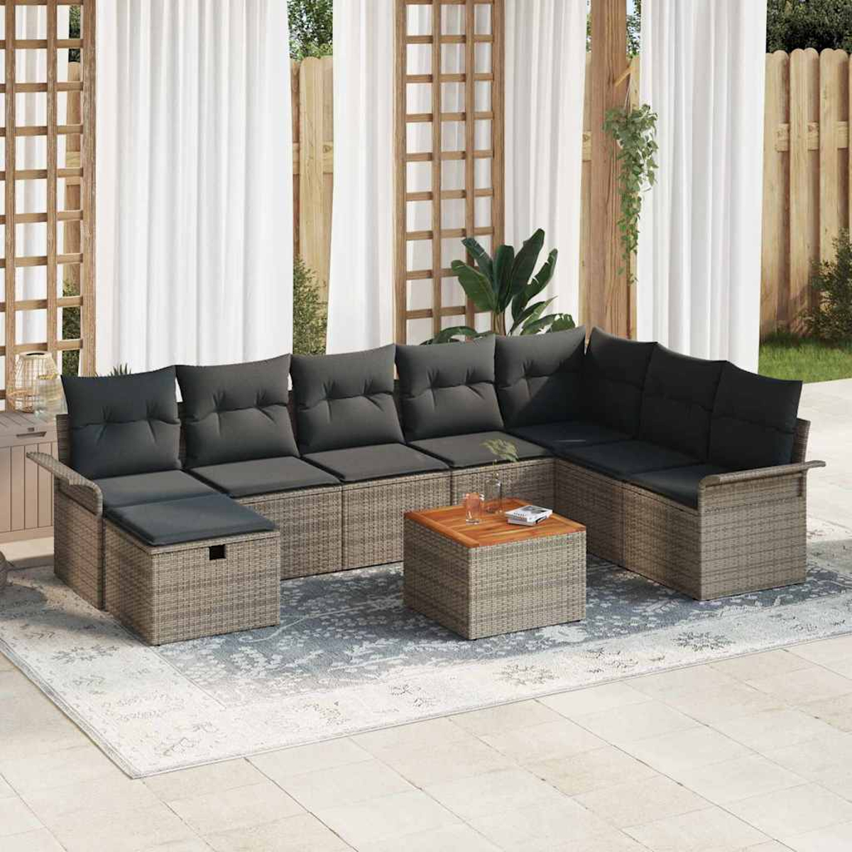 Ein Outdoor-Rattansofa mit schwarzen Kissen und einem Holztisch auf einem gemusterten Teppich. Transparente weiße Vorhänge und ein Holzzaun im Hintergrund.