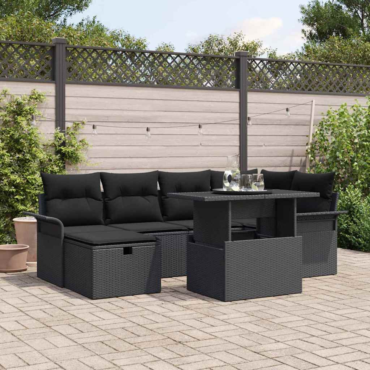 Juego de muebles de mimbre negro para exteriores con cojines y una mesa elevable, sobre un patio de ladrillos, con una valla al fondo.