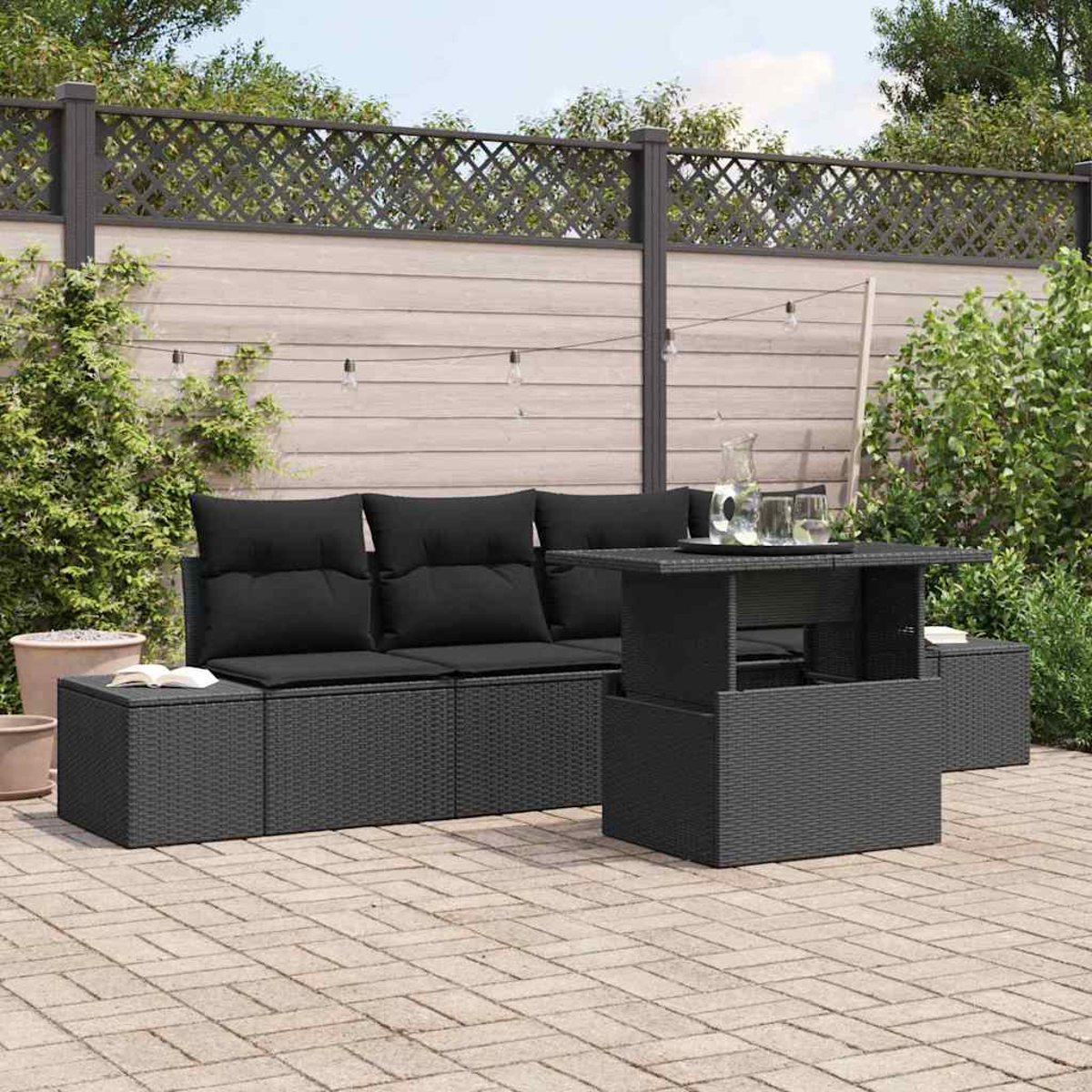 Garten-Terrassenset mit schwarzen Kissen und Tisch. Eine Glaskanne und Gläser stehen auf dem Tisch. Ziegelpflaster, Holzzaun im Hintergrund.