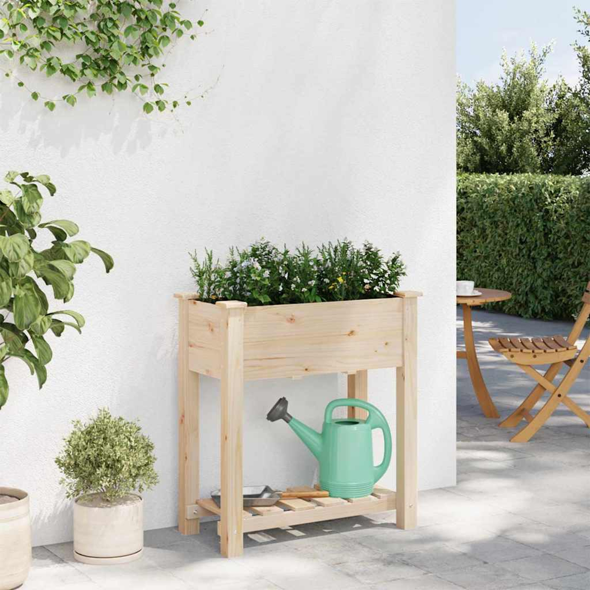 Fioriera in legno con piante, un annaffiatoio verde e un muro bianco. Sullo sfondo, un tavolo e una sedia all'aperto.