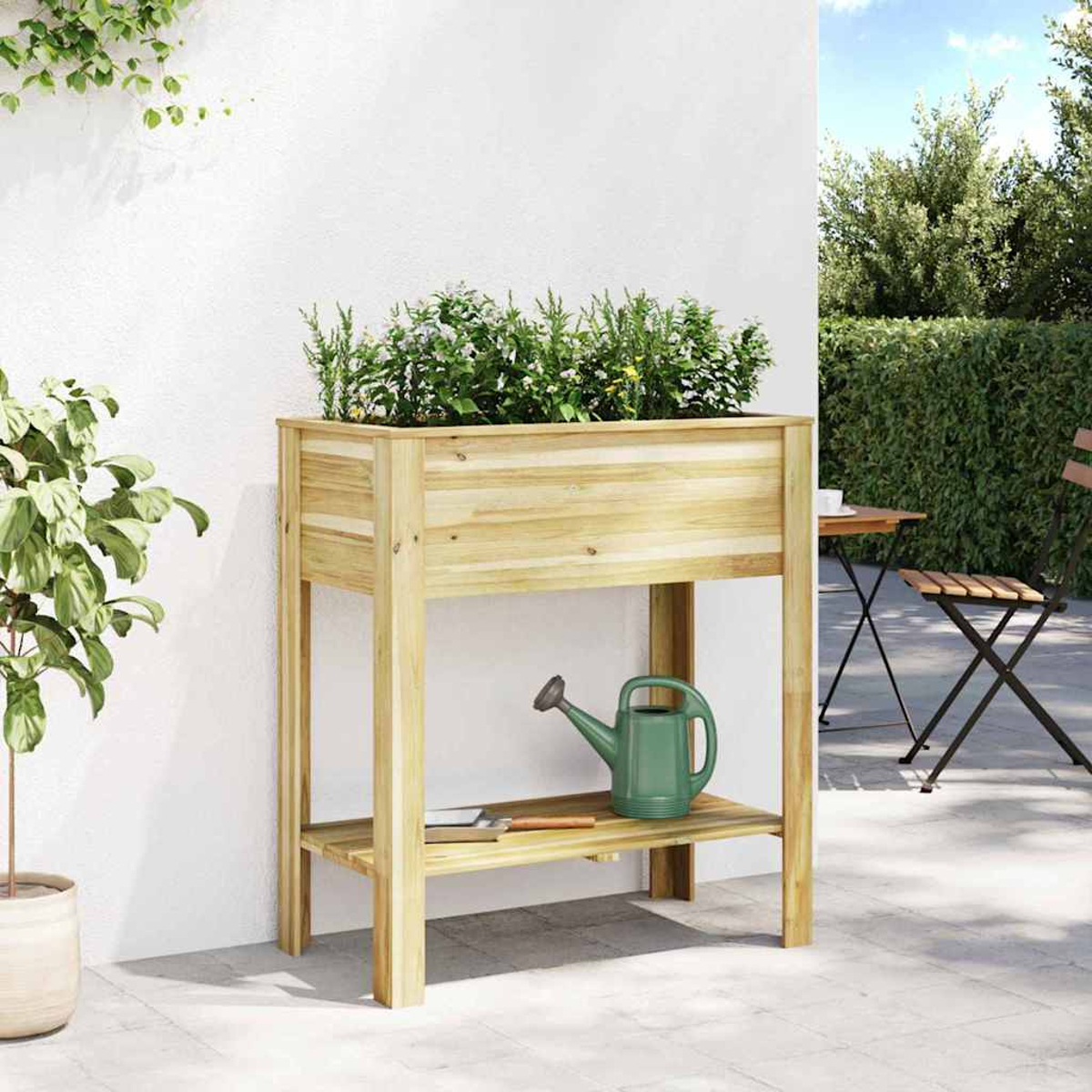 Fioriera in legno con piante. Un annaffiatoio verde è sullo scaffale sottostante. Parete bianca, ambiente esterno con tavolo e sedie.