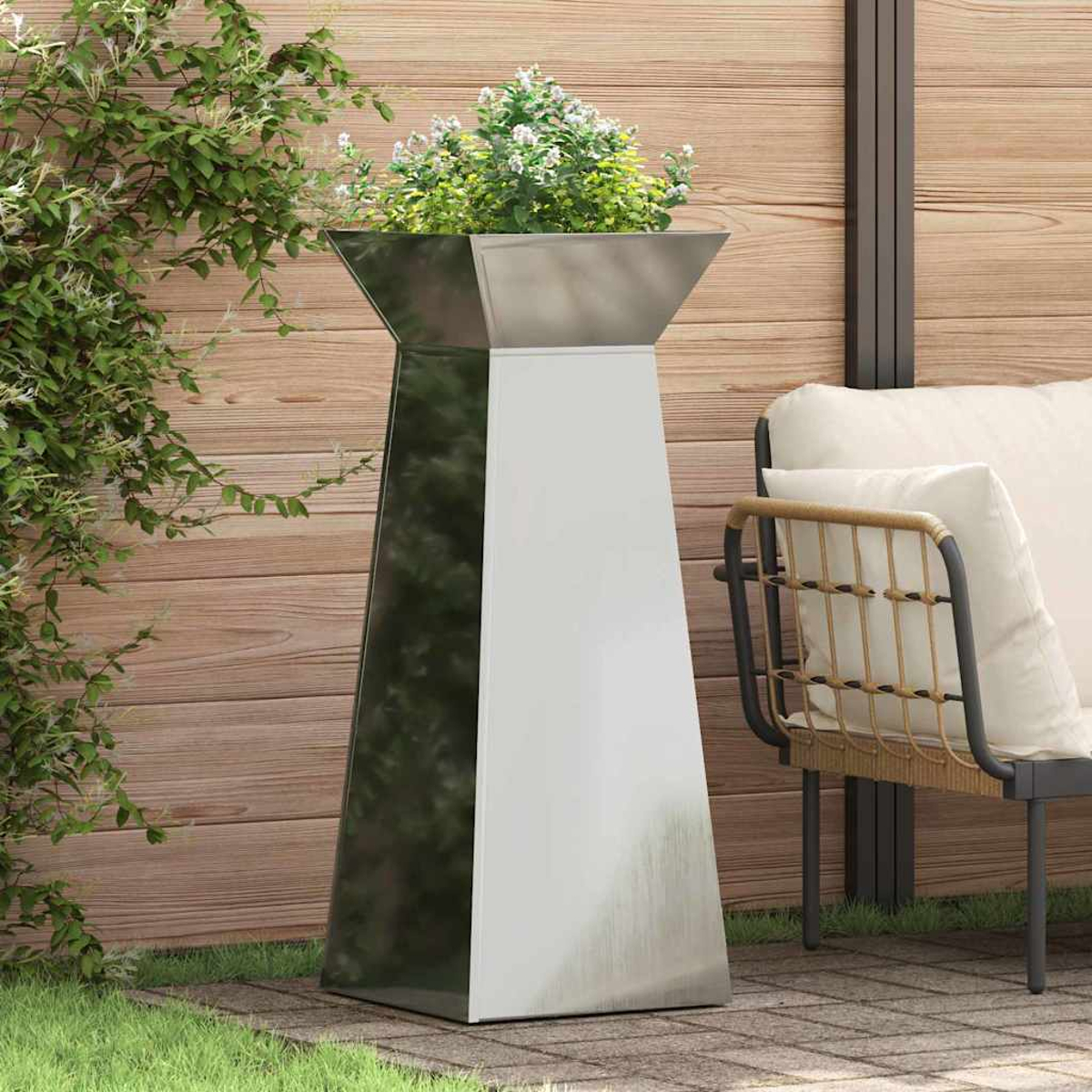 Un'alta e moderna fioriera con fiori si trova su un patio. Una sedia con cuscini è a destra. Il fogliame verde incornicia la scena.