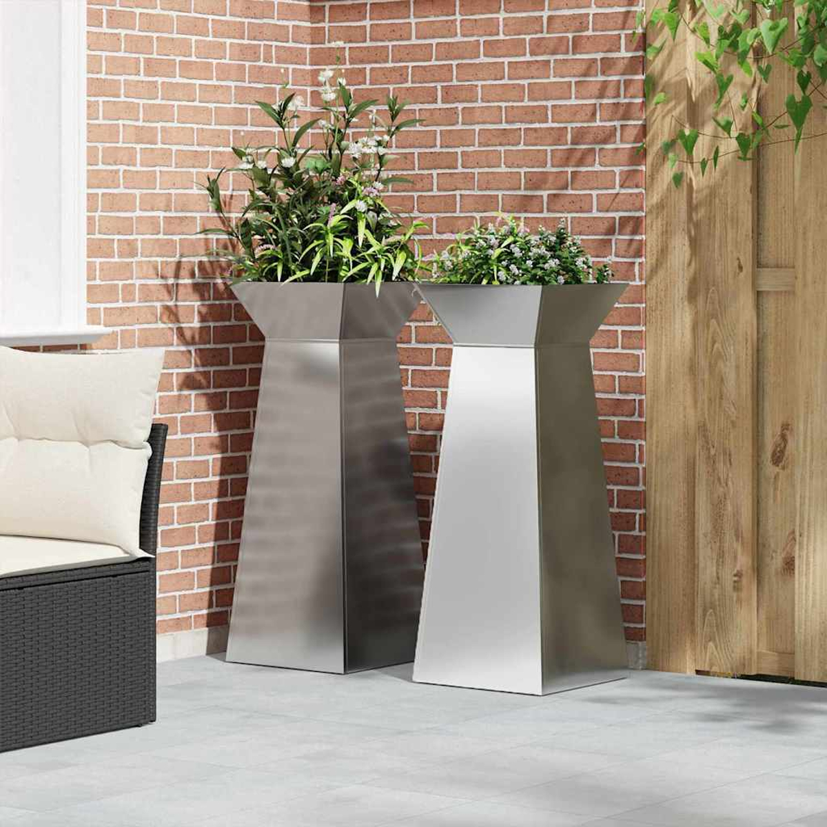 Due fioriere metalliche con piante su un patio. Un muro di mattoni e una recinzione di legno formano lo sfondo.