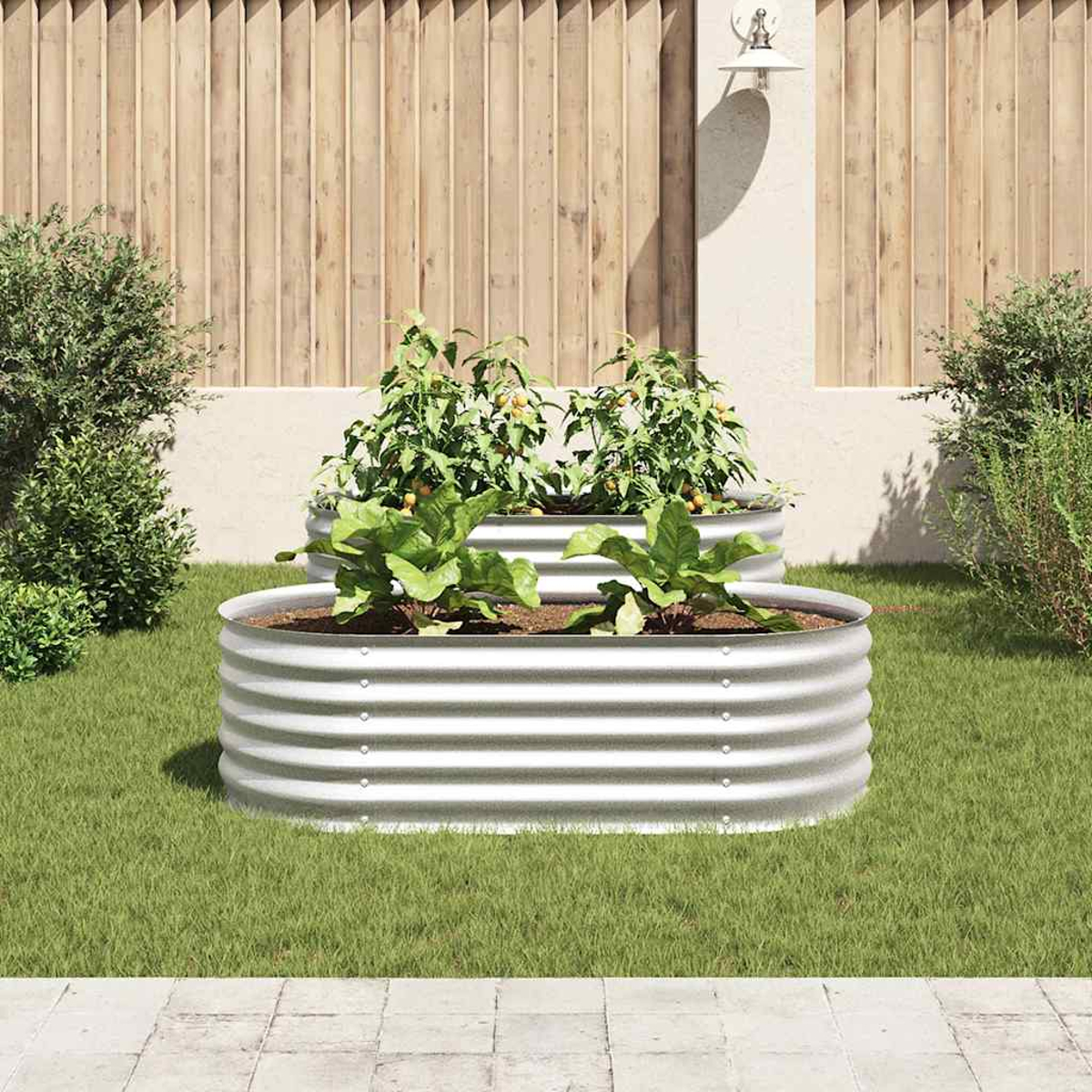 Zwei erhöhte Gartenbeete aus Metall mit Pflanzen. Der Hintergrund zeigt einen Holzzaun, grünes Gras und eine Wandleuchte.