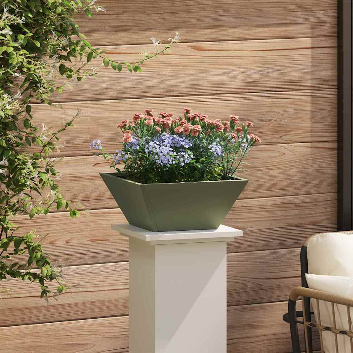 Una fioriera quadrata verde con fiori rosa e viola poggia su un piedistallo bianco. Parete in legno sullo sfondo.