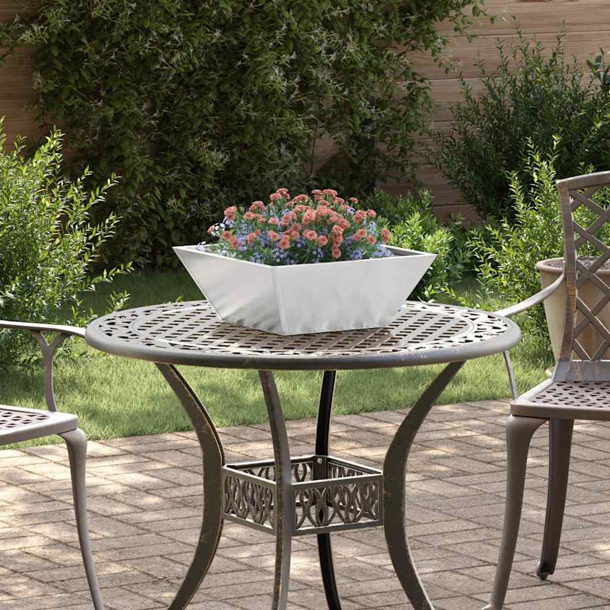 Una fioriera bianca con fiori è appoggiata su un tavolo di metallo. Il tavolo ha un design decorativo. Sedie circondano il tavolo su un patio di mattoni.