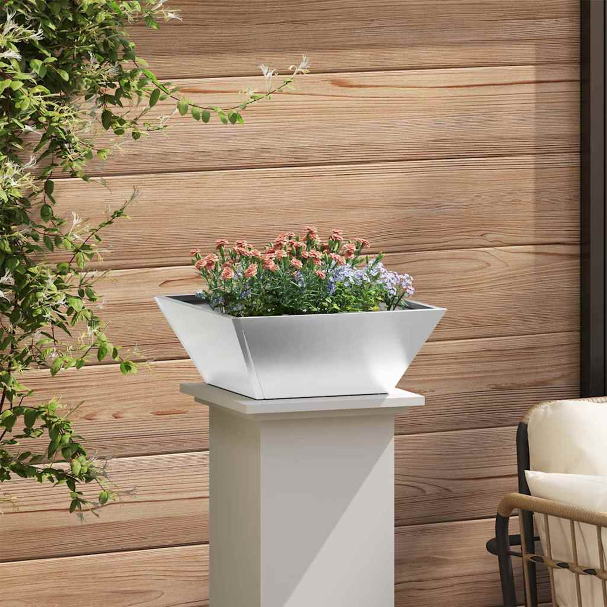 Una fioriera d'argento con fiori su un piedistallo bianco. Parete in legno sullo sfondo. Viti verdi a sinistra e sedia a destra.