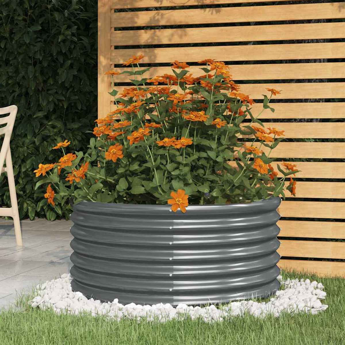 Ein runder, grauer Metallpflanzer mit orangefarbenen Blumen steht auf weißen Steinen. Der Hintergrund zeigt einen Holzzaun und Grün.