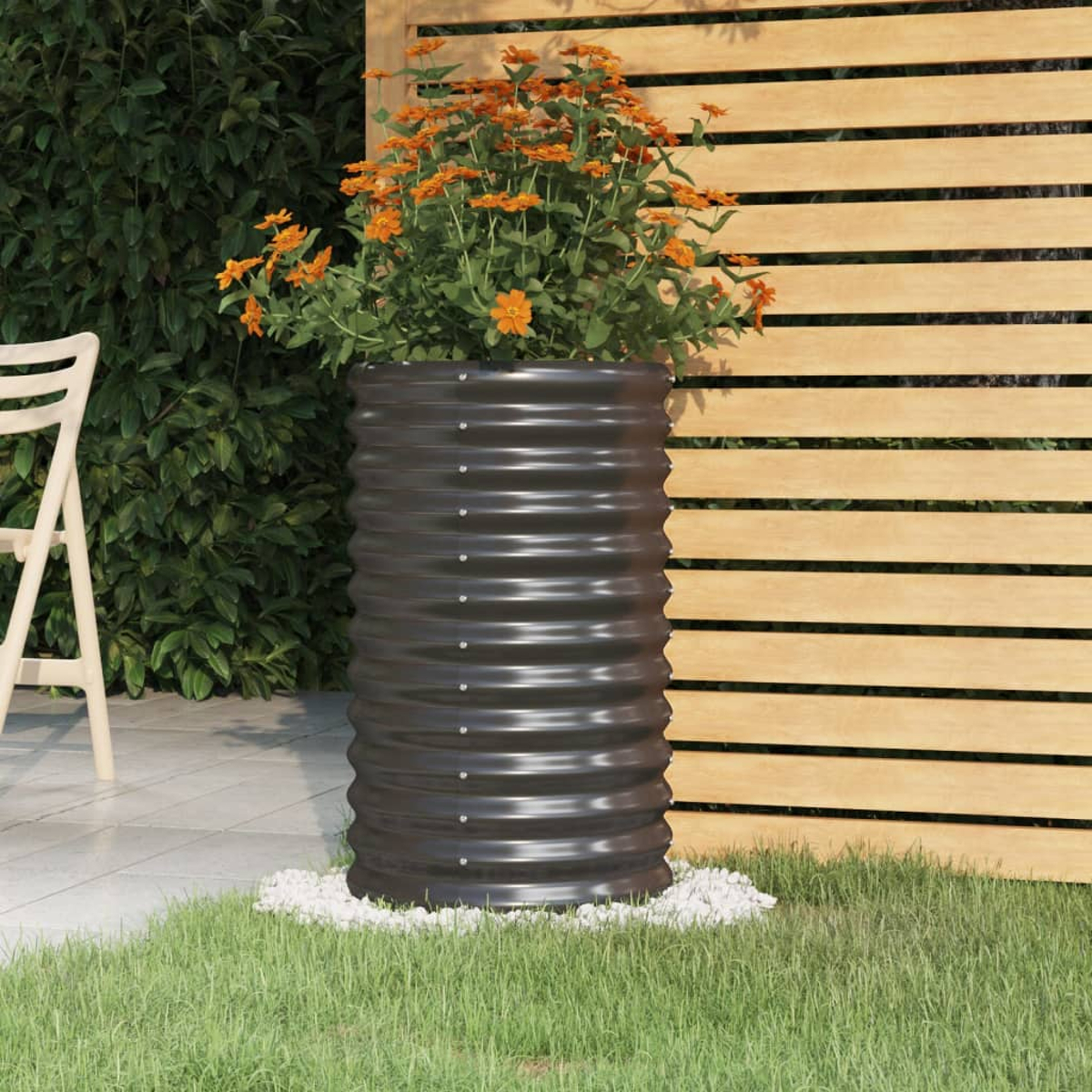 Ein dunkler Metallpflanzkübel mit orangefarbenen Blumen steht auf weißen Steinen. Ein Holzzaun und grünes Laub bilden den Hintergrund.