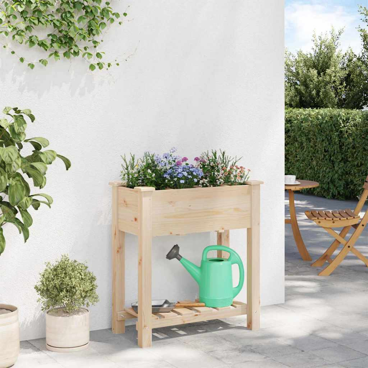 Fioriera in legno con fiori, un annaffiatoio verde e piccole piante su un patio. Un tavolo e una sedia sono visibili sullo sfondo.