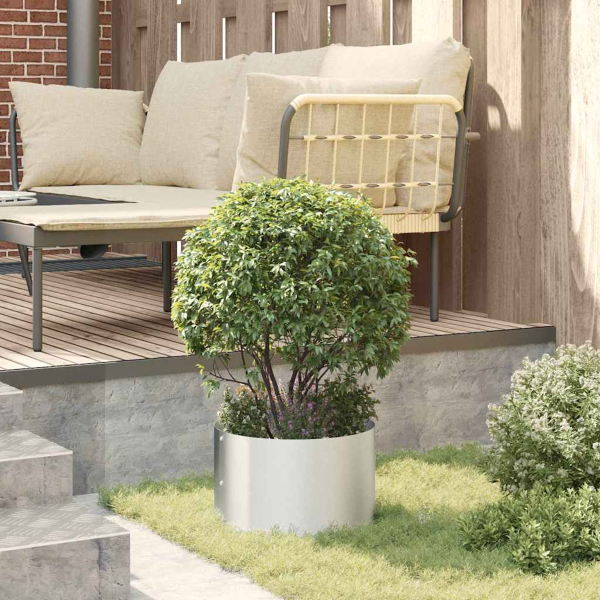 Eine Terrasse mit einem grünen Busch in einem Metallpflanzgefäß. Ein beiges Sofa und Holzzaun im Hintergrund. Grünes Gras im Vordergrund.
