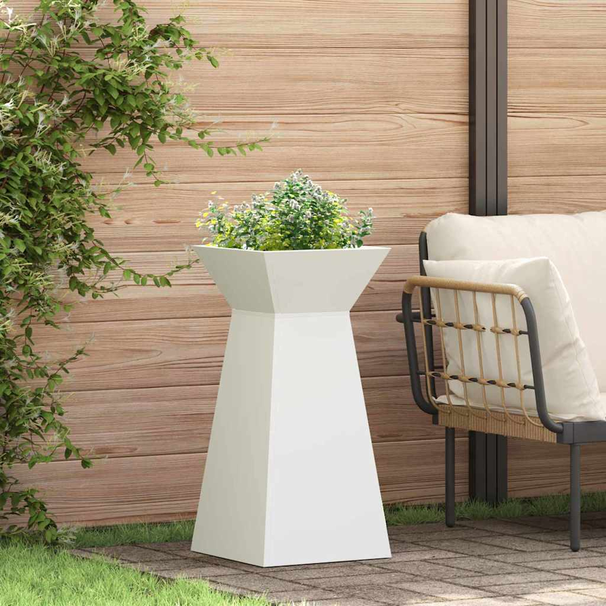 Un portavaso bianco con piante verdi. Una sedia a destra. Sfondo con recinzione in legno e fogliame verde.