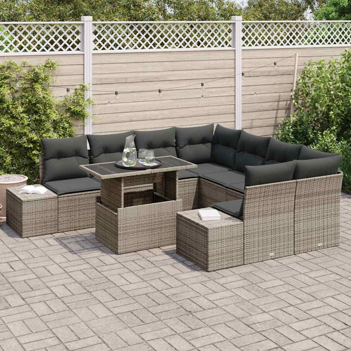 Gartenmöbel-Set. Korbsessel mit grauen Kissen, ein Tisch mit Getränken und eine Backstein-Terrasse. Zaun und Grün im Hintergrund.