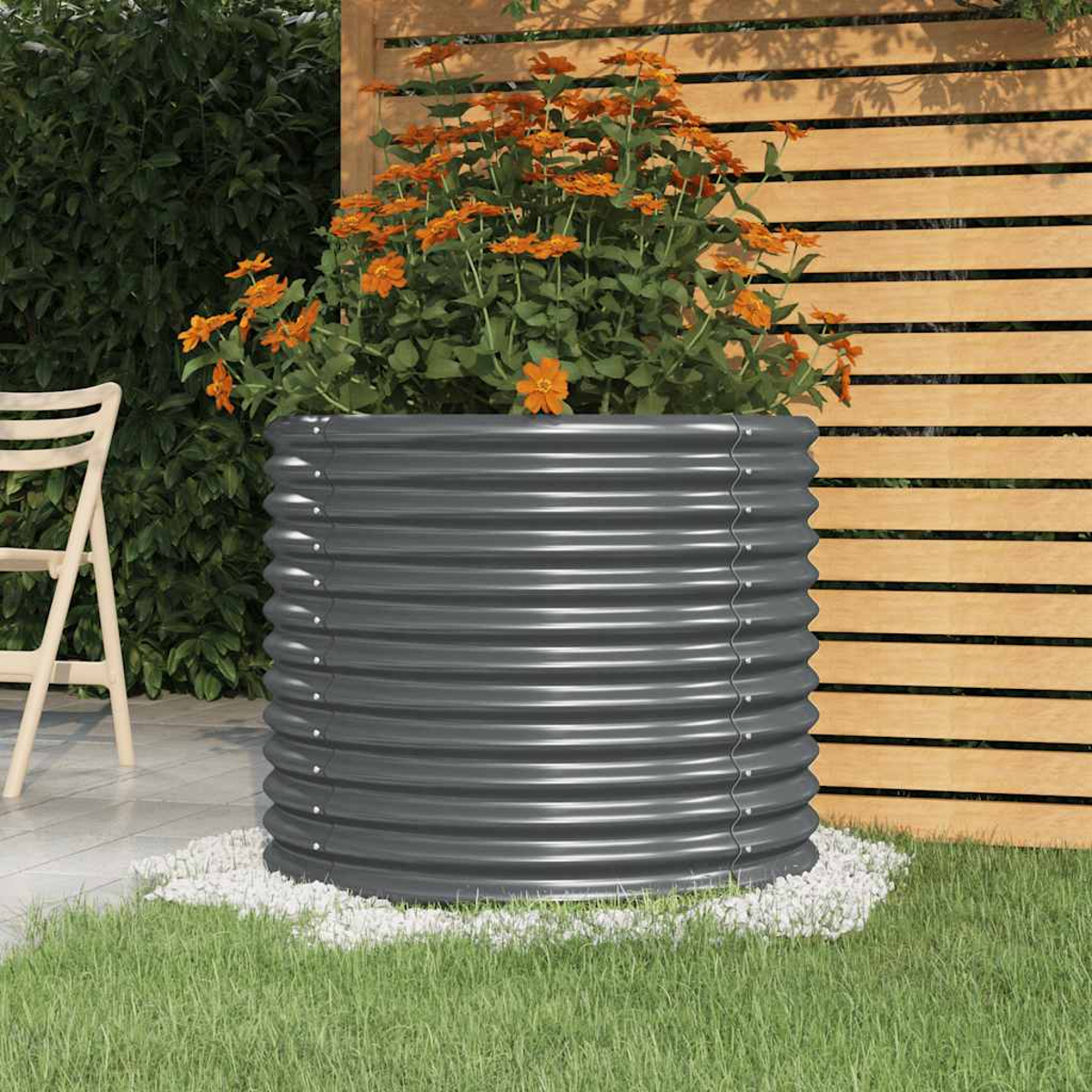 Ein grauer Metallpflanzer mit orangefarbenen Blumen steht auf weißen Steinen. Ein Holzzaun und grüner Rasen sind im Hintergrund.