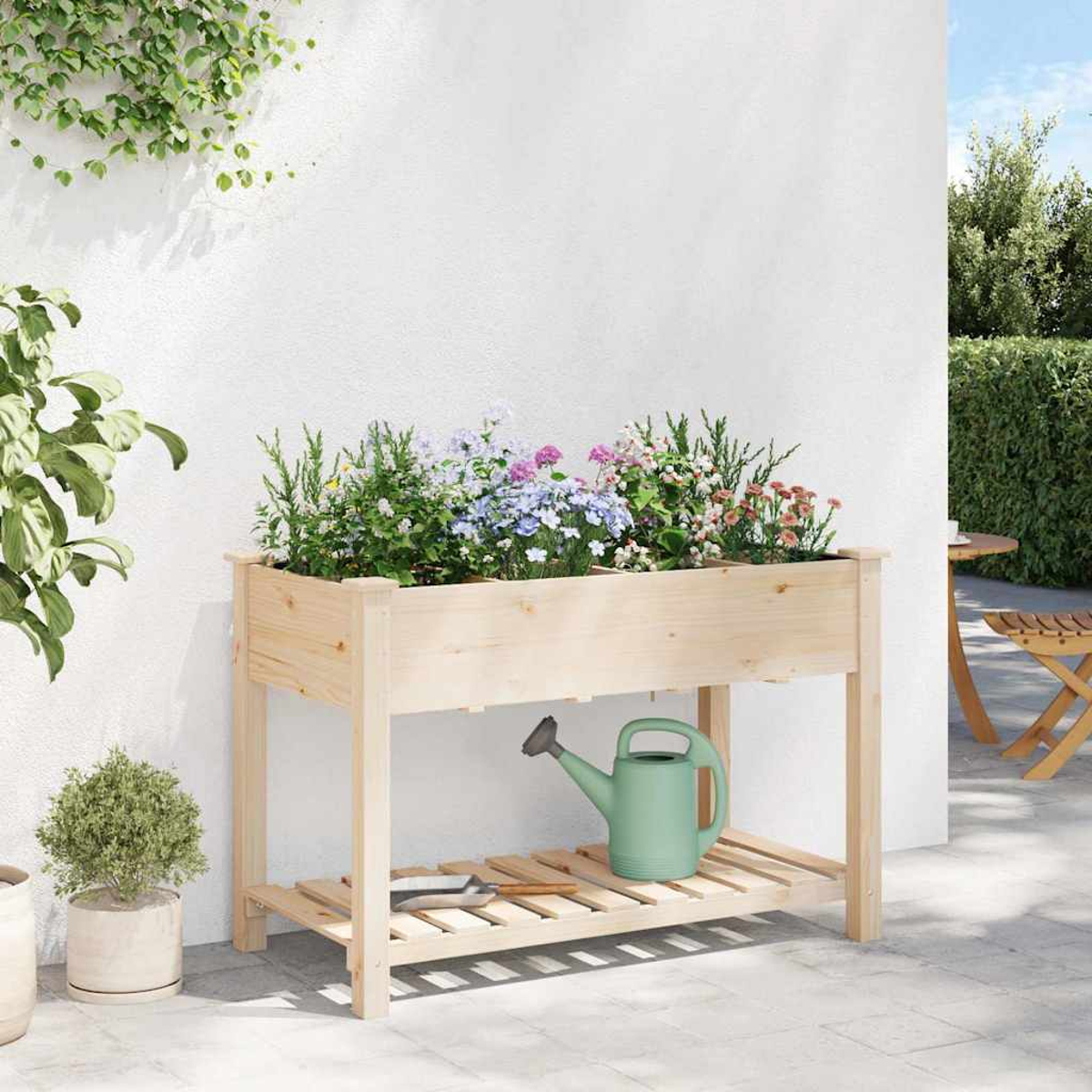 Holz-Pflanzkasten mit bunten Blumen, einer grünen Gießkanne und einer kleinen Pflanze. Weißer Wandhintergrund.
