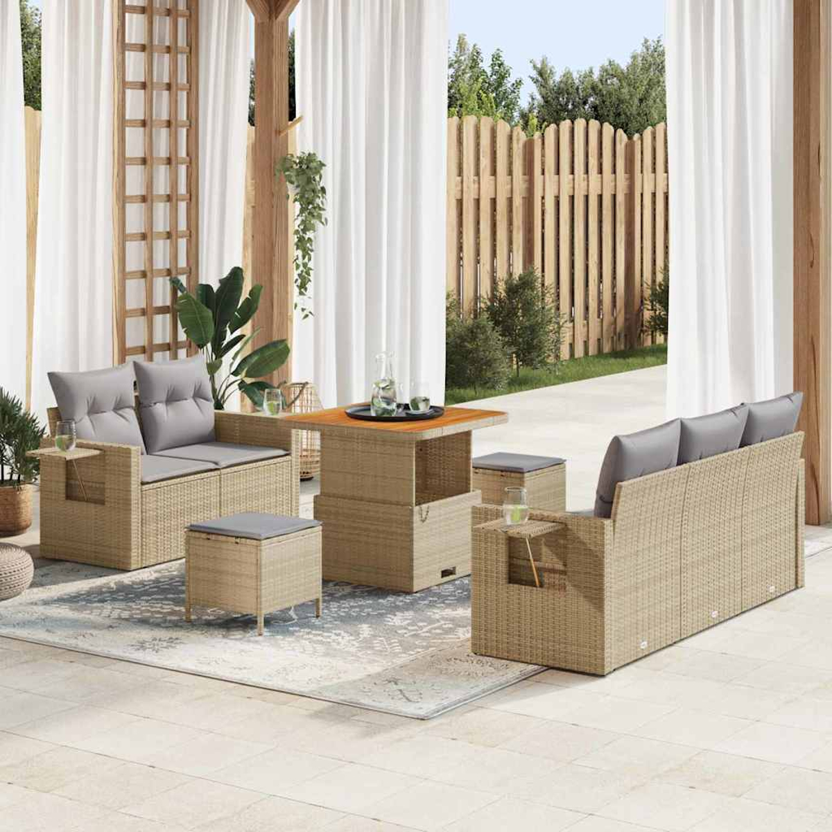 Terrassenset mit Sofa, Tisch und Ottomanen. Graue Kissen auf beigem Korbgeflecht. Vorhänge, Holzzaun und Pflanzen im Hintergrund.