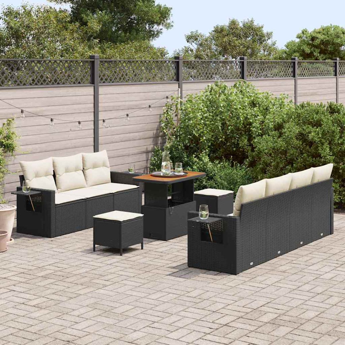 Outdoor-Terrassenset: Schwarze Korbsessel, Tisch und Ottomanen mit cremefarbenen Kissen, angeordnet auf einer Backsteinterrasse. Ein Zaun und Grün sind im Hintergrund.