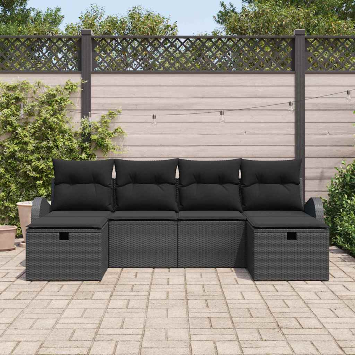 Graues Rattan-Sofa im Freien mit schwarzen Kissen. Dahinter befindet sich ein Holzzaun und Grün. Die Szene spielt sich auf einer Terrasse ab.