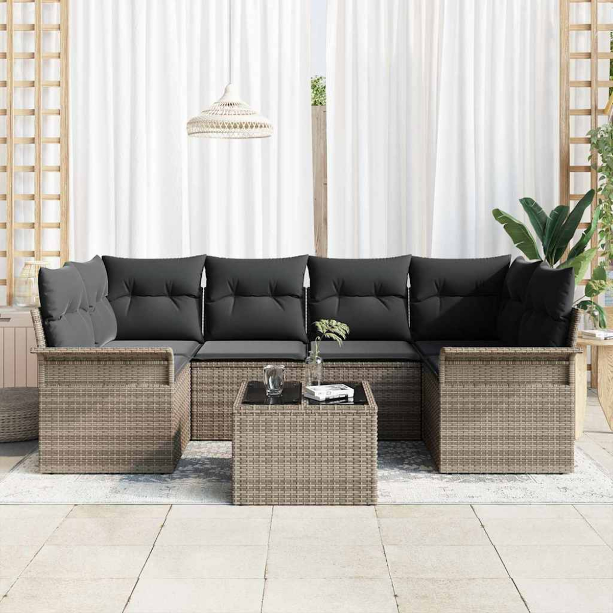 Ein Outdoor-Sofa aus Rattan mit dunklen Kissen und einem Tisch mit Glasplatte. Weiße Vorhänge und eine gewebte Lampe sind im Hintergrund.