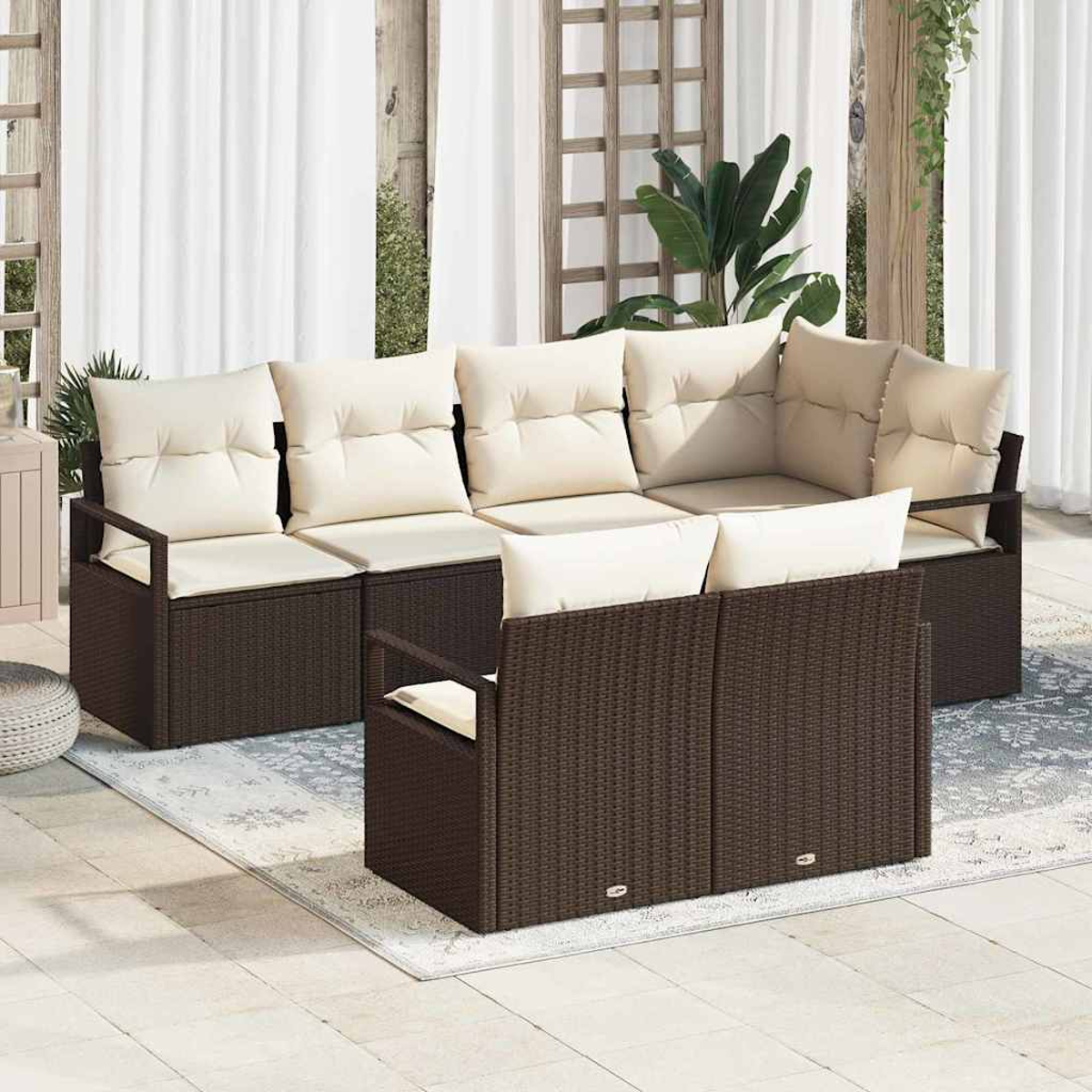 Gartenmöbel-Set aus Korbgeflecht mit cremefarbenen Kissen auf einer Terrasse. Weiße Vorhänge und Pflanzen im Hintergrund.