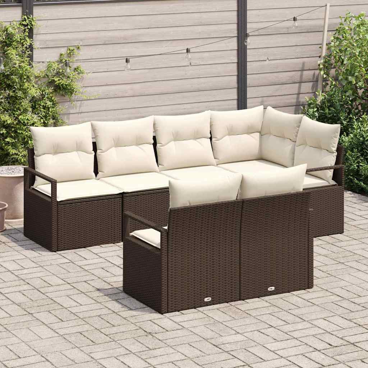 Outdoor-Rattan-Ecksofa mit cremefarbenen Kissen. Auf einer Steinterrasse, mit Holzzaun im Hintergrund. Lichterketten sind aufgehängt.