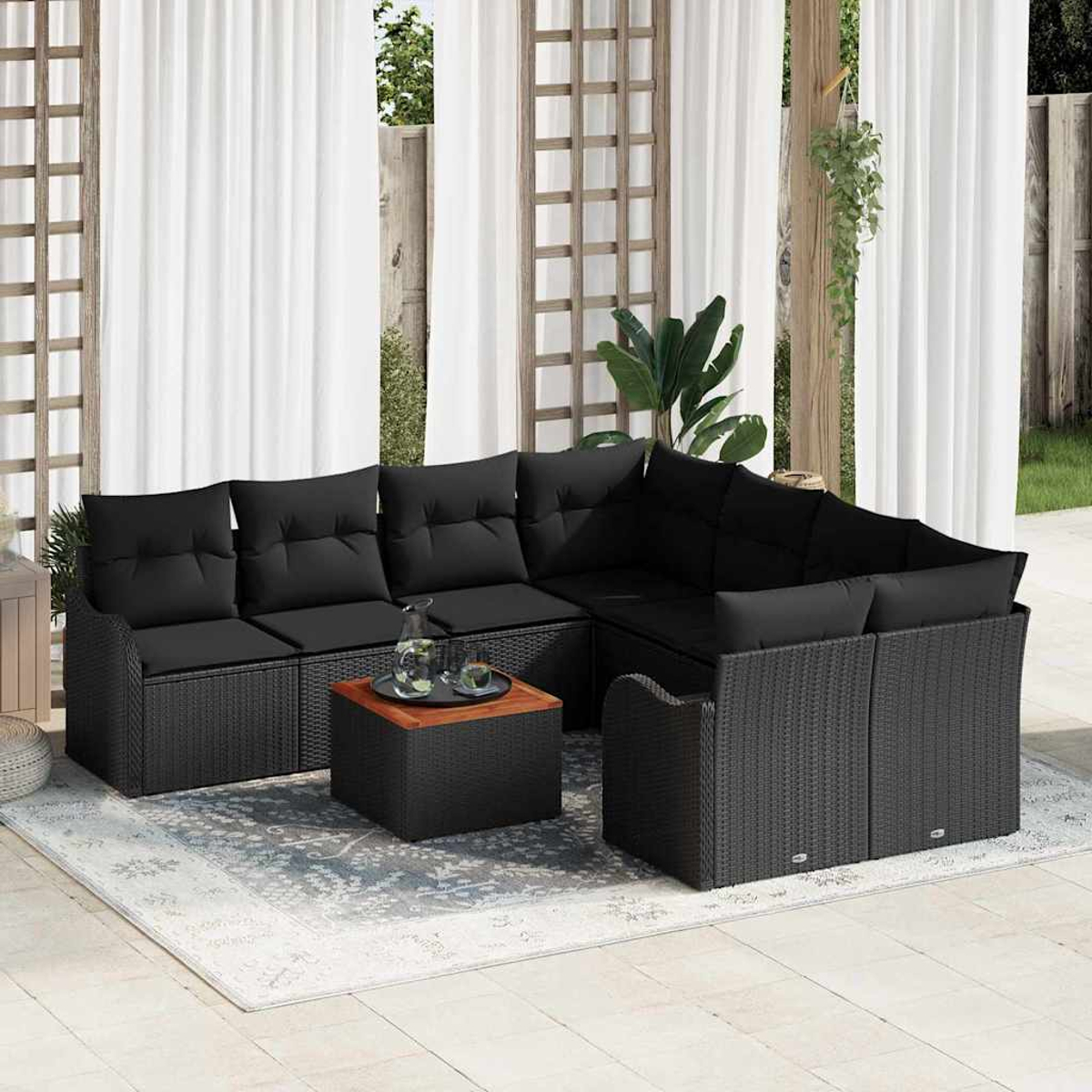Sofá seccional para exteriores con cojines negros y base tejida, una mesa pequeña y una alfombra en un patio con cortinas blancas.