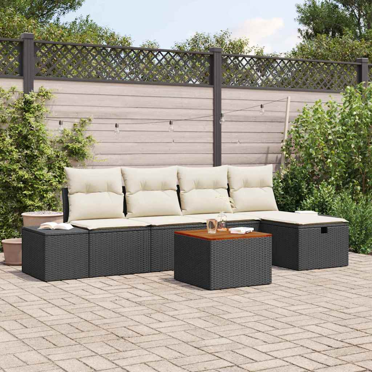 Juego de patio exterior con mimbre gris y cojines beige. Incluye sofá, dos otomanas y mesa de centro. Fondo: valla y vegetación.