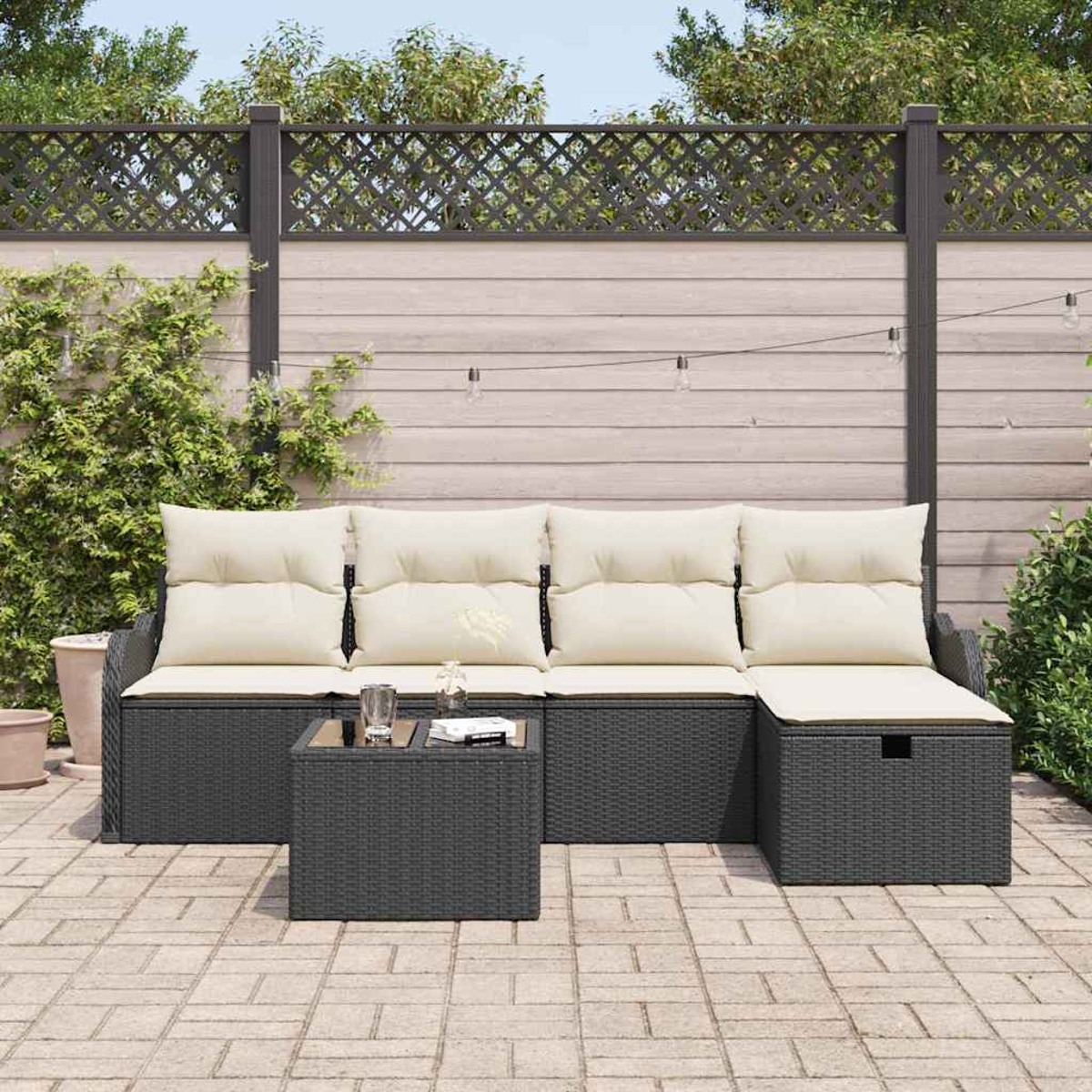 Garten-Terrassenset mit dunkelgrauem Korbsessel, cremefarbenen Kissen und einem passenden quadratischen Tisch. Vor Holzzaun und Grün.
