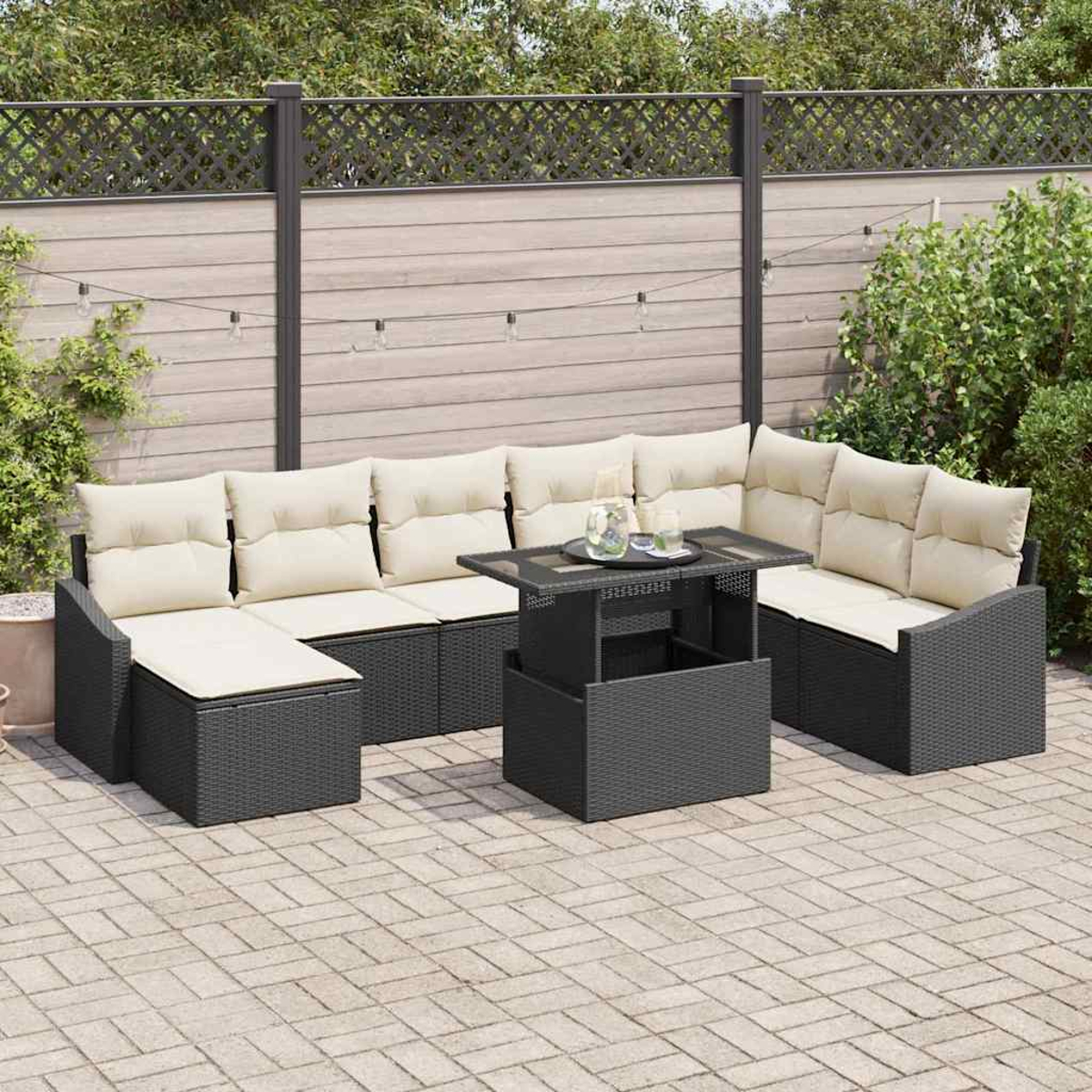 Outdoor-Sitzgruppe mit beigefarbenen Kissen, schwarzen Korbmöbeln und einem Tisch mit Hubfunktion auf einer Terrasse. Hintergrund: Zaun, Pflanzen.
