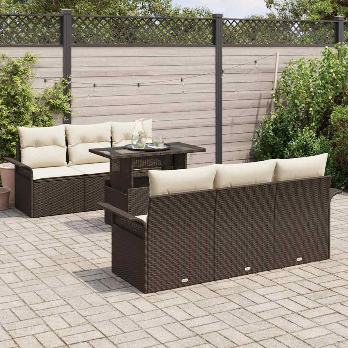 Garten-Set mit braunen Korbmöbeln, cremefarbenen Kissen und einem Tisch mit Glasplatte. Vor einem Holzzaun und Grün.