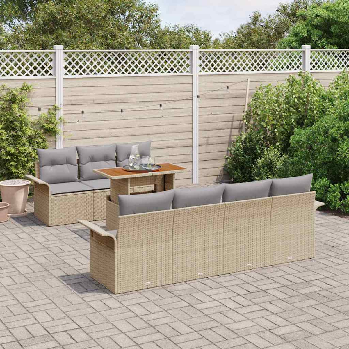 Ein Outdoor-Sitzmöbel-Set aus Korbgeflecht mit grauen Kissen und einem kleinen Tisch steht auf einer Backstein-Terrasse. Im Hintergrund ist ein weißer Zaun zu sehen.