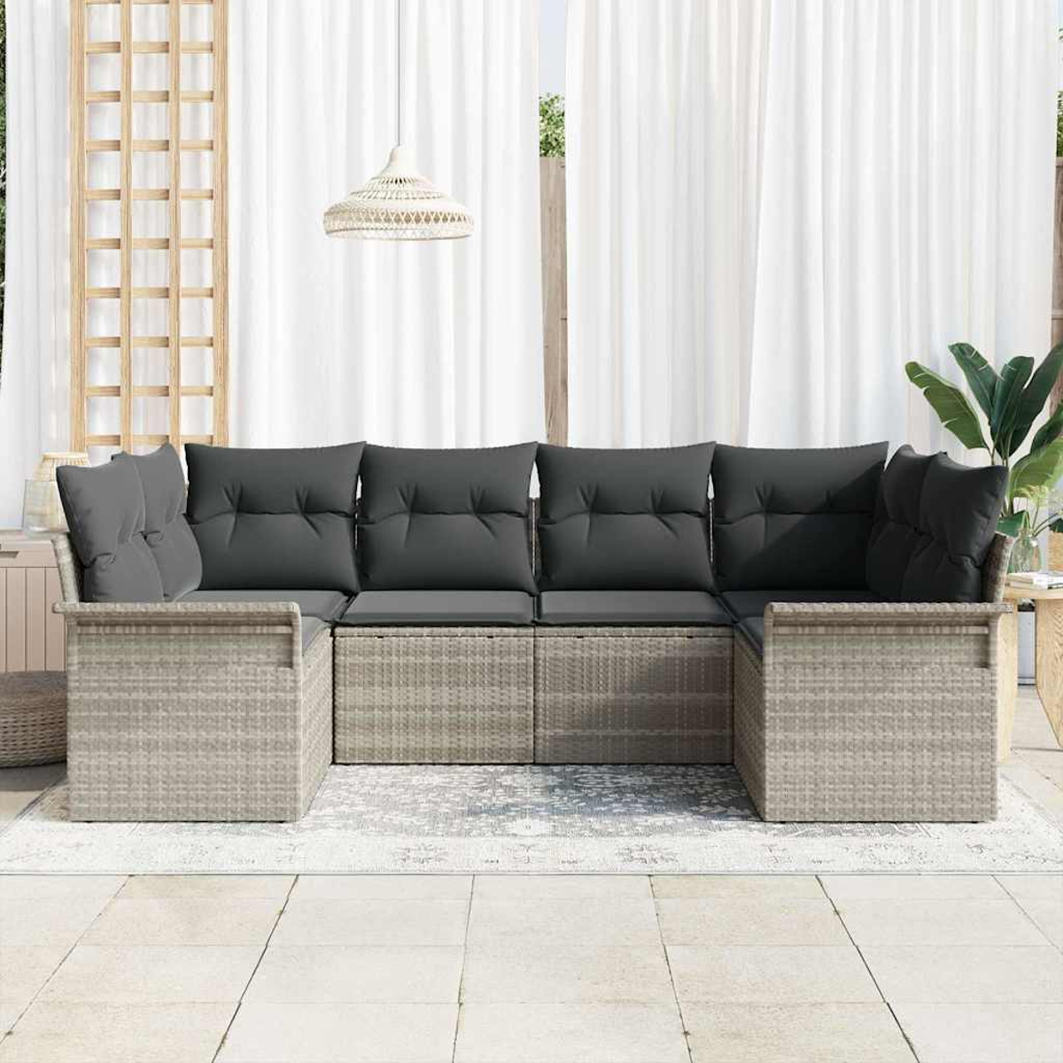 Graues Rattansofa mit grauen Kissen steht auf einem Teppich. Weiße Vorhänge sind dahinter. Eine Leuchte hängt oben.