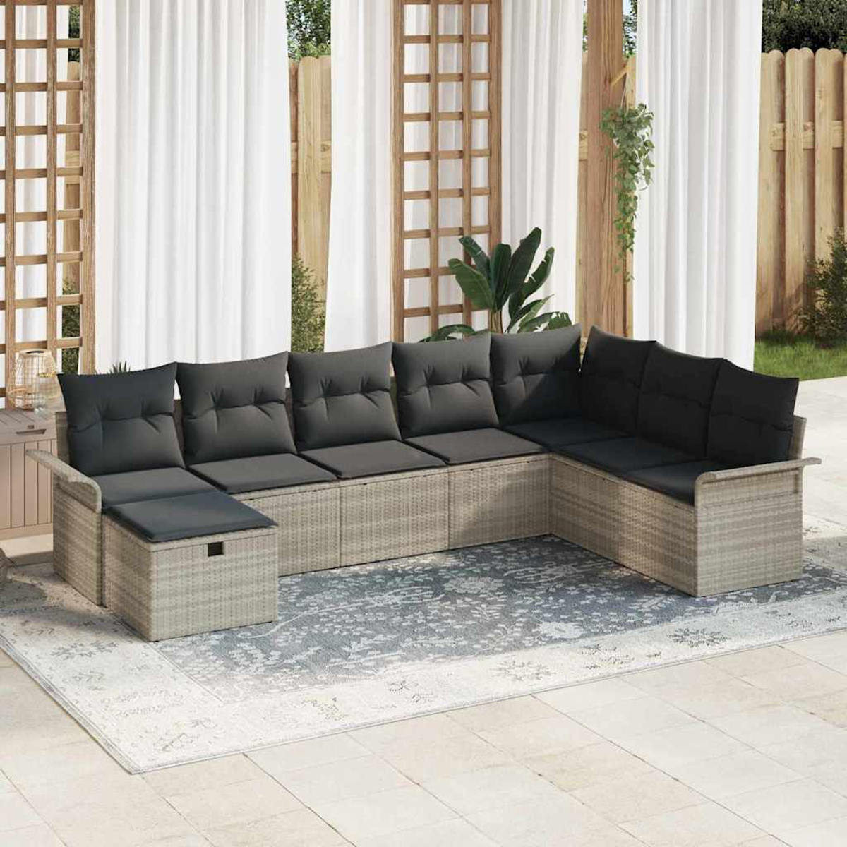 Ein graues Ecksofa aus Korbgeflecht mit dunkelgrauen Kissen steht auf einem gemusterten Teppich. Dahinter sind weiße Vorhänge und ein Holzzaun.