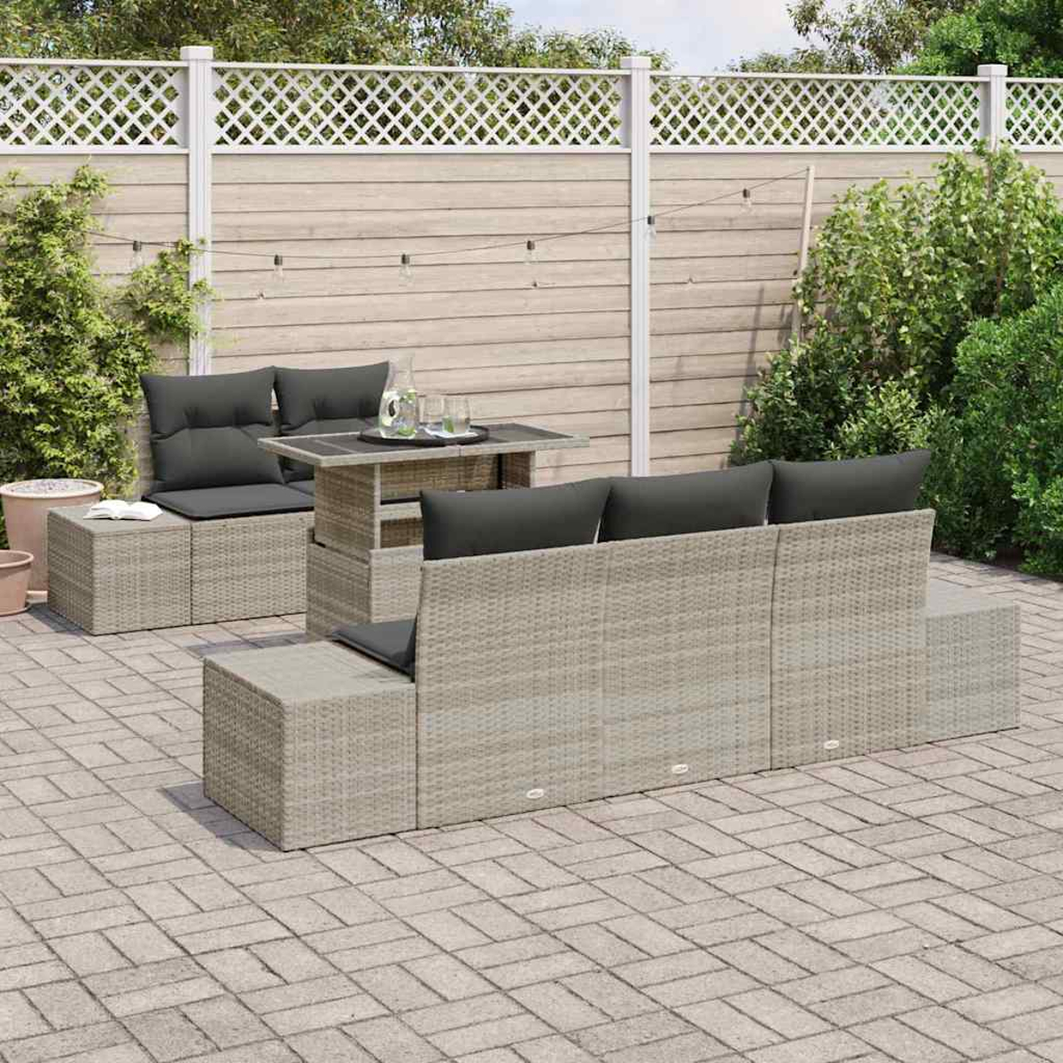 Eine Terrasse mit Rattanstühlen, grauen Kissen und einem Tisch. Im Hintergrund ein Holzzaun und Grün. Gepflasterter Boden.