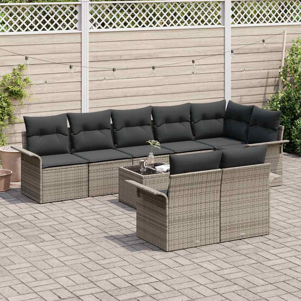 Gartenmöbel-Set mit grauen Kissen. Graue Korbmöbel auf einer gepflasterten Terrasse. Hintergrund zeigt einen Holzzaun und Grün.
