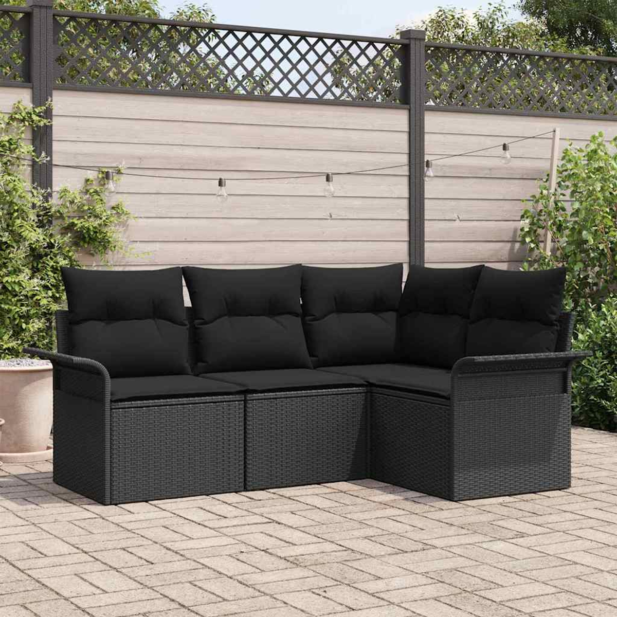 Ein schwarzes Korbsessel-Ecksofa mit schwarzen Kissen steht auf einer Ziegelstein-Terrasse. Ein Holzzaun und Lichterketten befinden sich im Hintergrund.