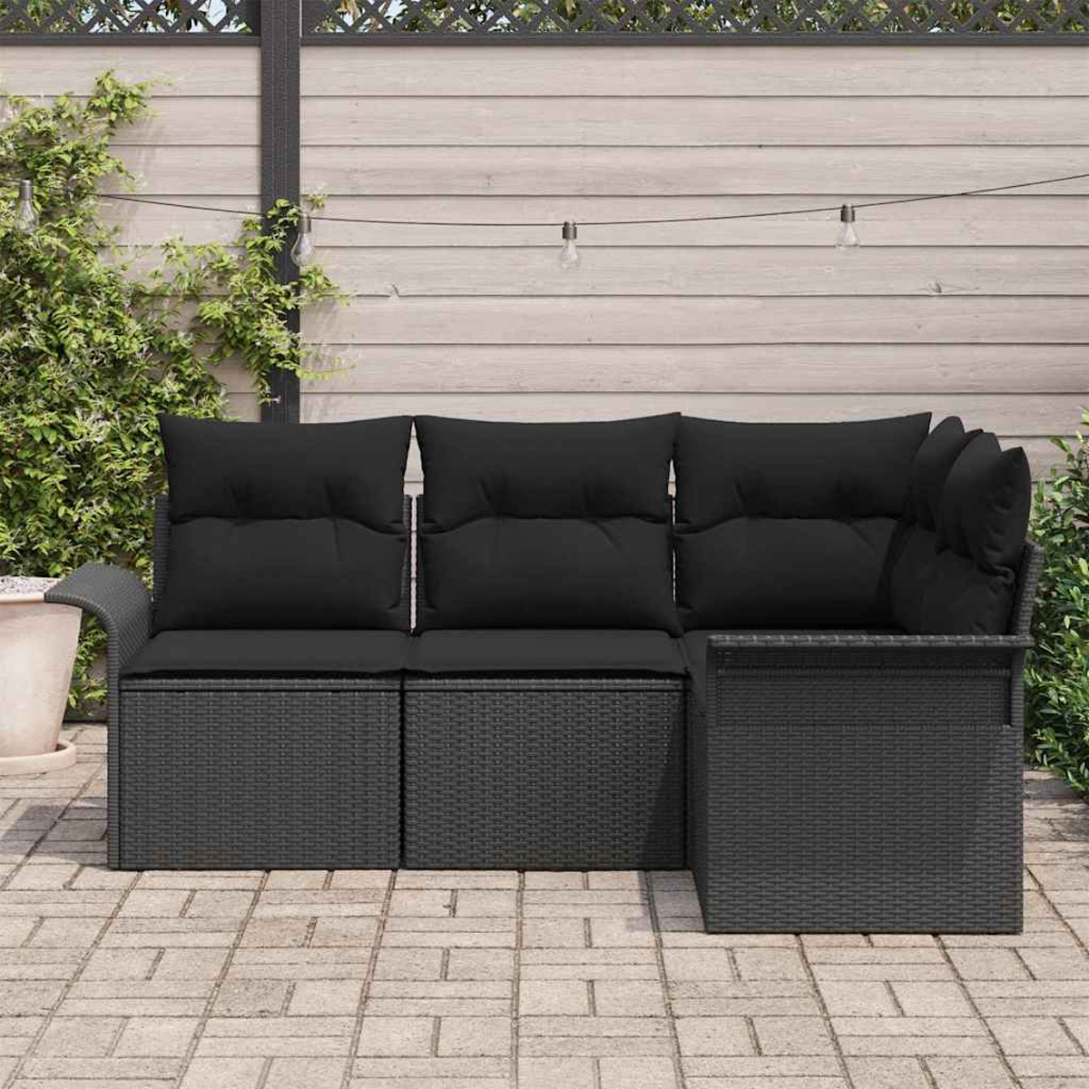 Ein schwarzes Outdoor-Sofa aus Korbgeflecht mit schwarzen Kissen steht auf einer Ziegelsteinterrasse. Ein Holzzaun und Grünzeug sind im Hintergrund.