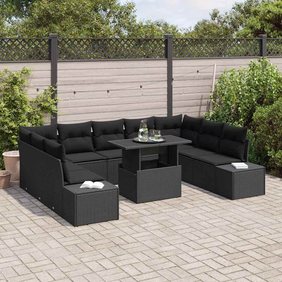 Garten-Sitzgruppe mit schwarzen Kissen, Tisch mit Getränken, auf Steinterrasse, mit Holzzaun im Hintergrund.