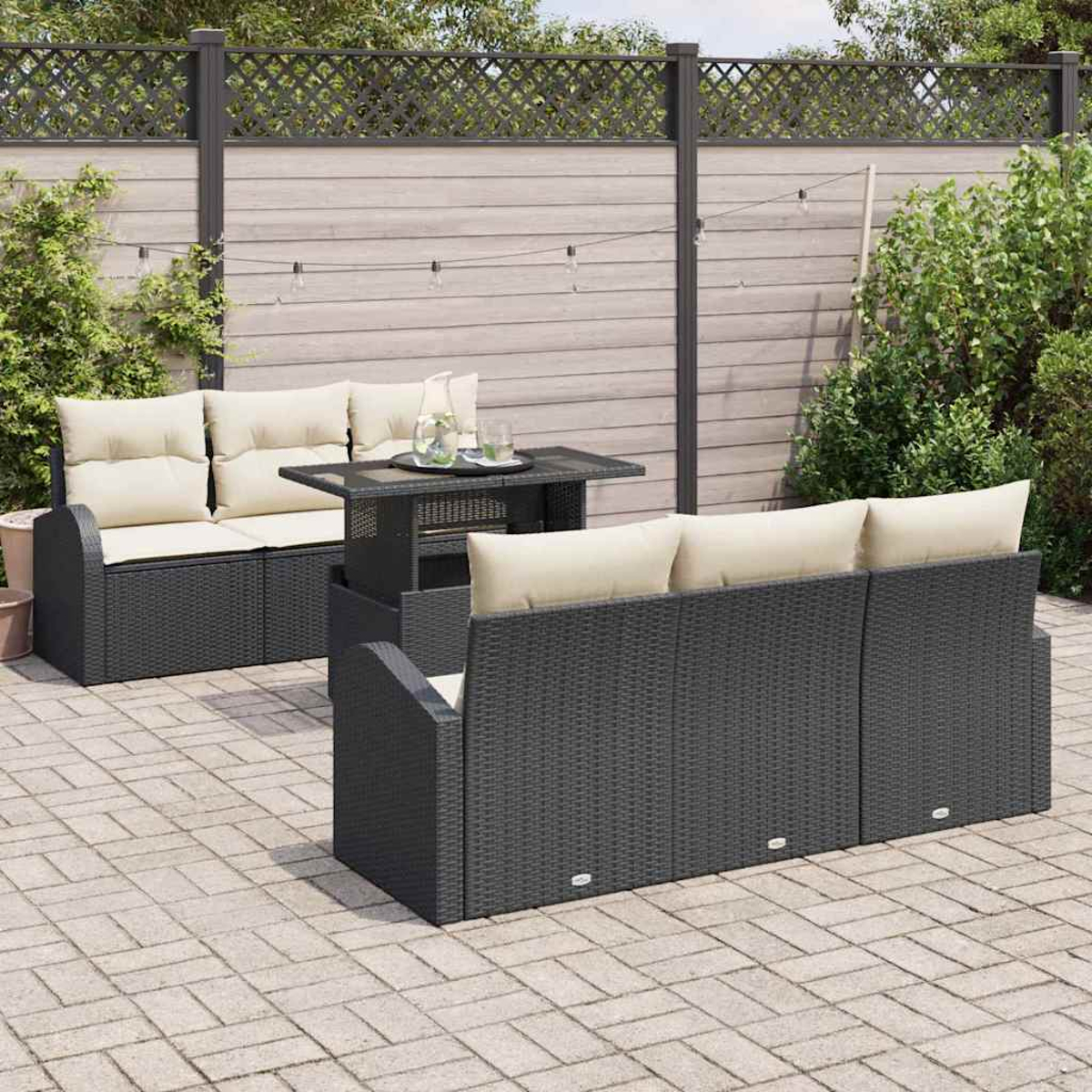 Gartenmöbel-Set: graue Korbmöbel mit beigefarbenen Kissen, kleiner Tisch und gepflasterte Terrasse. Holzzaun im Hintergrund.